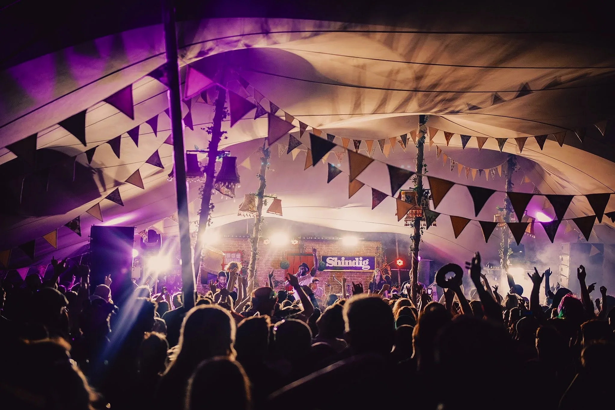 Crowd at a music festival with a stage, decorated with bunting under a large tent, illuminated by purple and white lights.