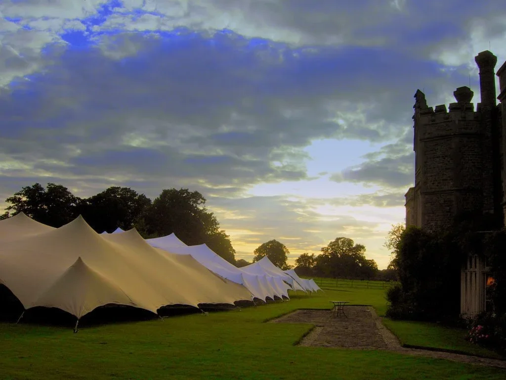 Row of white tents on a grassy field near a historic stone castle, with a cloudy sky at sunset.