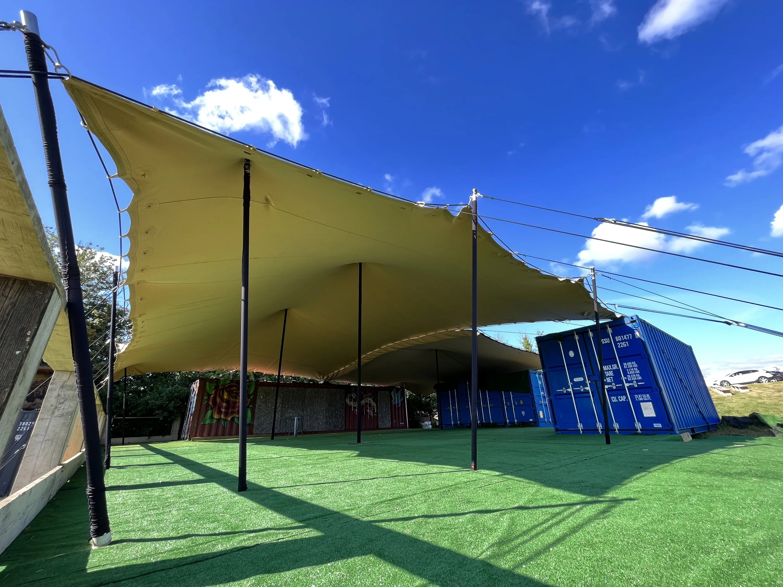 A yellow fabric canopy stretched overhead, supported by black poles, shading an outdoor area with green artificial turf, next to a wooden fence and large shipping containers.