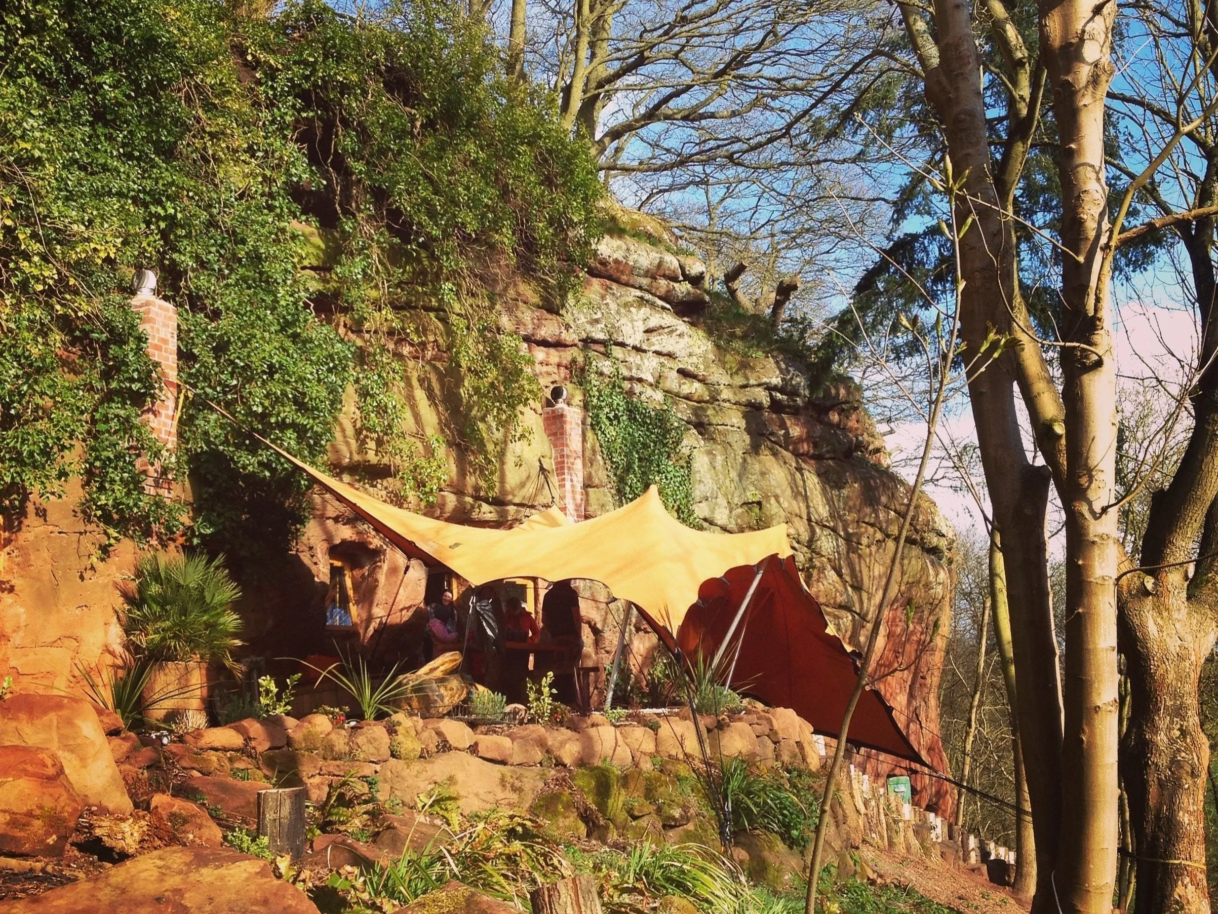 Outdoor scene with trees, rocky cliff, orange canopy tents, and some people sitting underneath in a park.