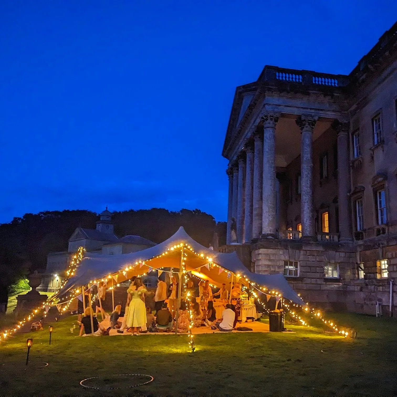 An evening outdoor event with a lit tent surrounded by string lights, people gathering, and a historic building with columns in the background under a dark blue sky.