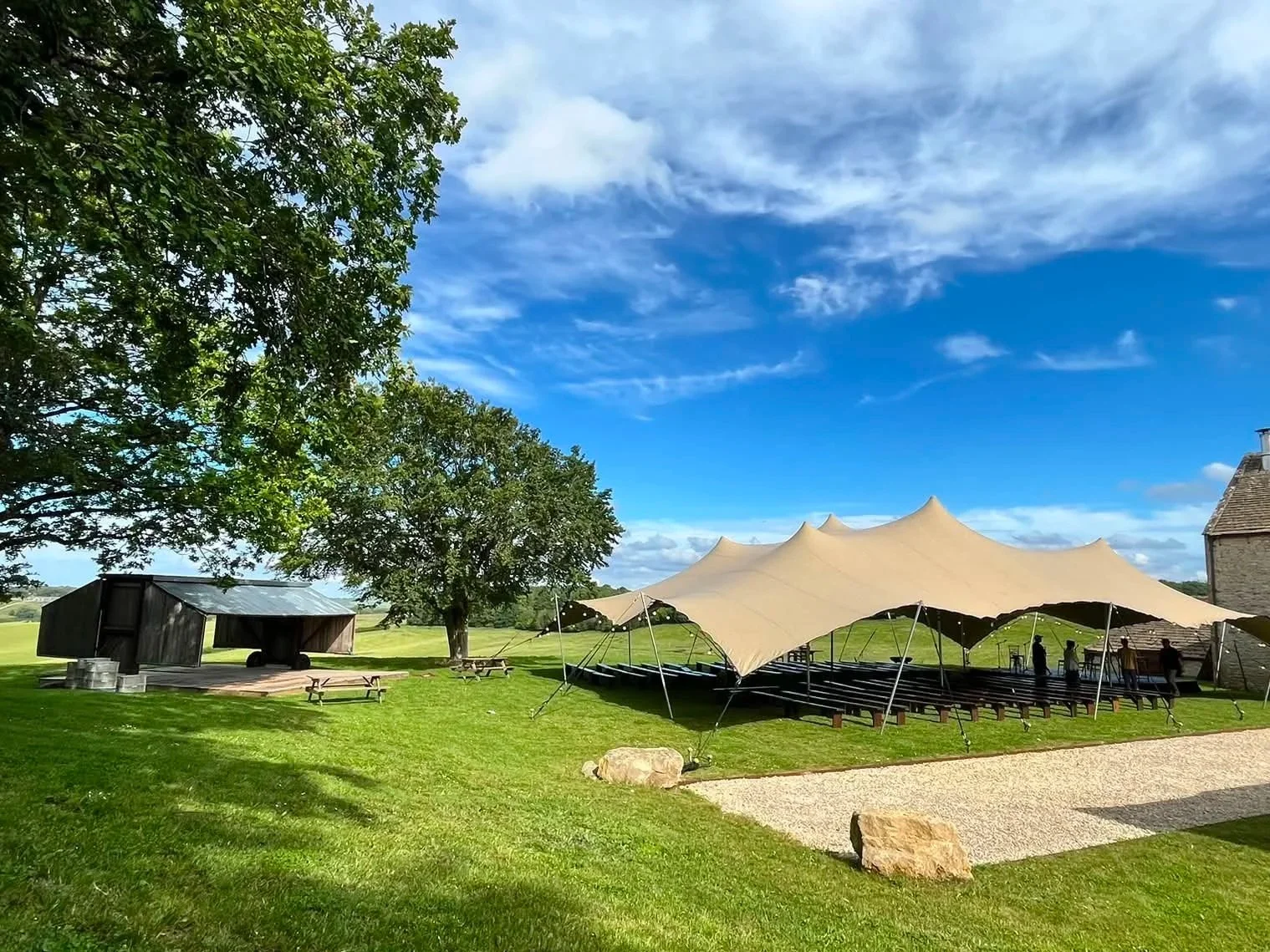 Outdoor event setup with a large beige tent canopy, stage with people, black wooden shed, picnic tables, trees, and a grassy field under a partly cloudy sky.
