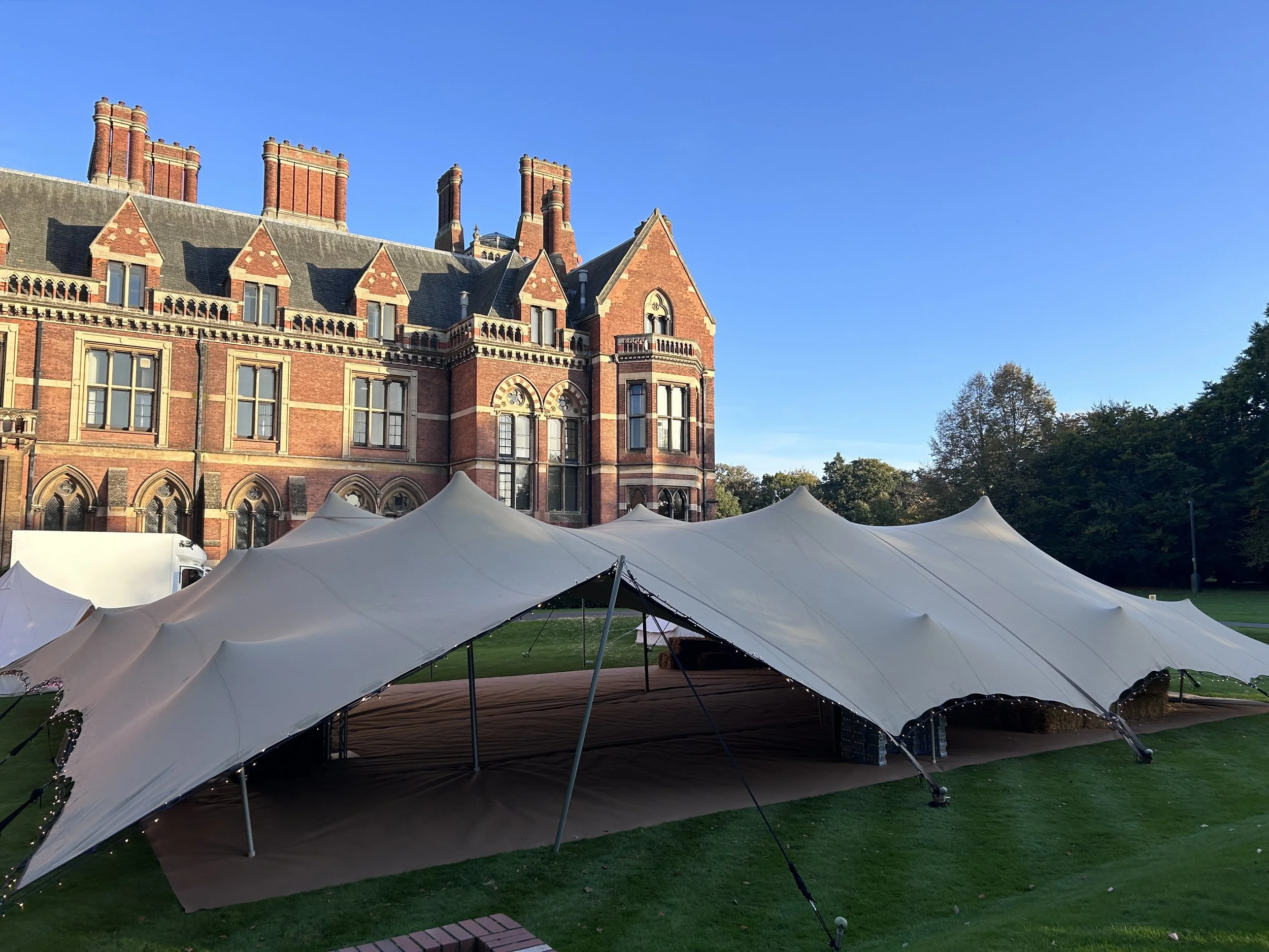 A large historic building with red brick walls, multiple chimney stacks, and ornate windows. In front of the building, there is a large outdoor event tent with lighting, set up on a grassy area next to a brick walkway under a clear blue sky.