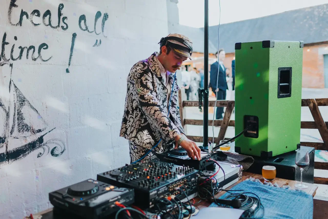 A DJ wearing a patterned shirt and a cap, focusing on playing music at an outdoor event, with DJ equipment, a large green speaker, and drinks on the table.