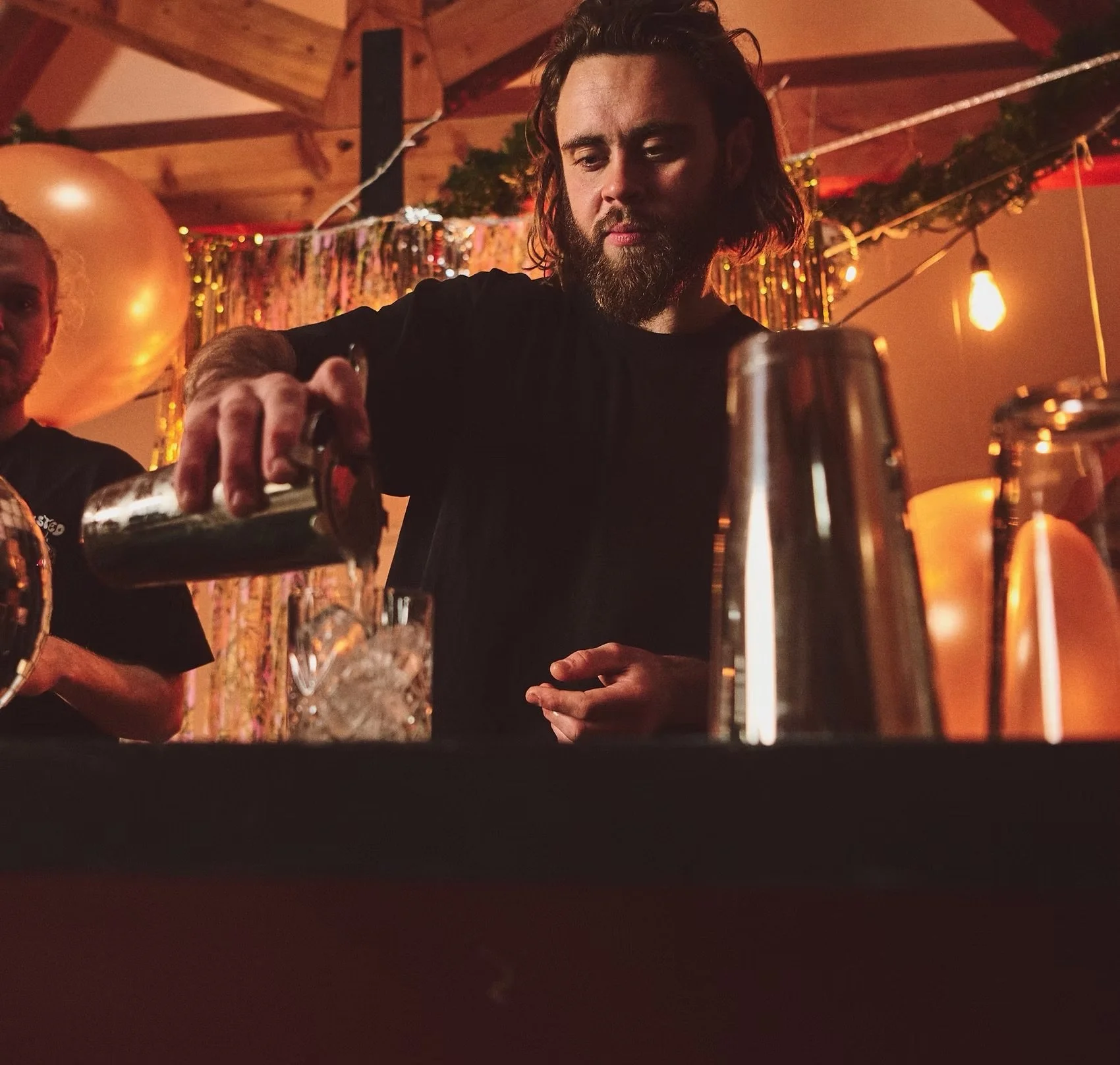 A man with long hair and a beard serving drinks at a party or gathering. The room has warm lighting, wooden ceiling, and festive decorations, including a large balloon, tinsel, and hanging light bulbs.
