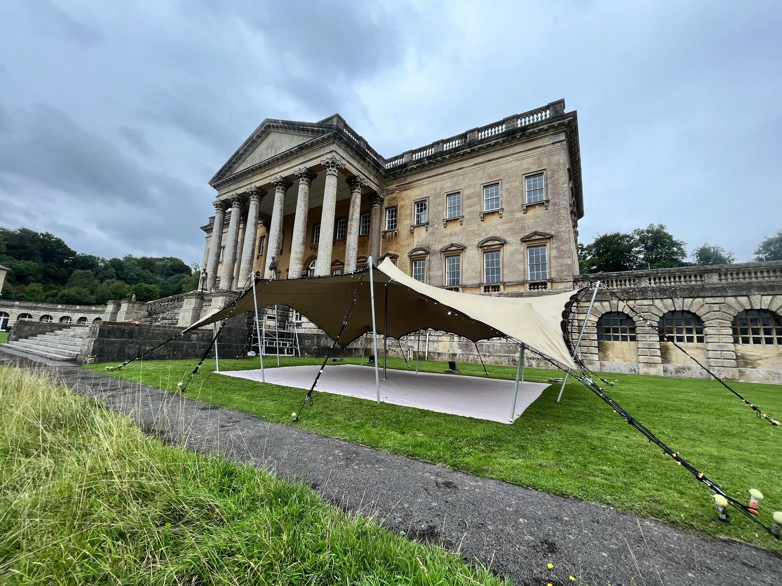 A large classical-style building with tall columns and a triangular pediment. There is a landscaped area with grass and a tent-like structure in front. The sky is partly cloudy.