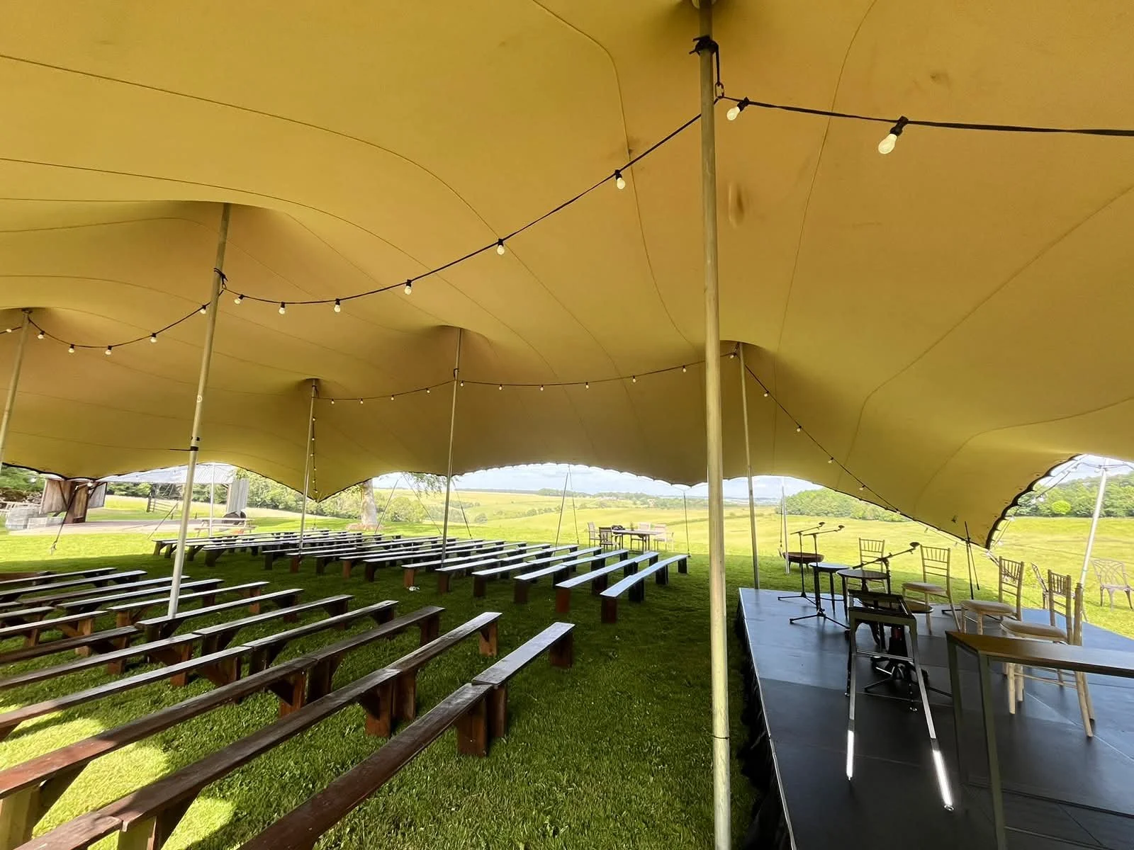 Empty outdoor event space under a large yellow tent with string lights, wooden benches, and a stage with chairs and microphones, set in a grassy field with trees and open landscape.