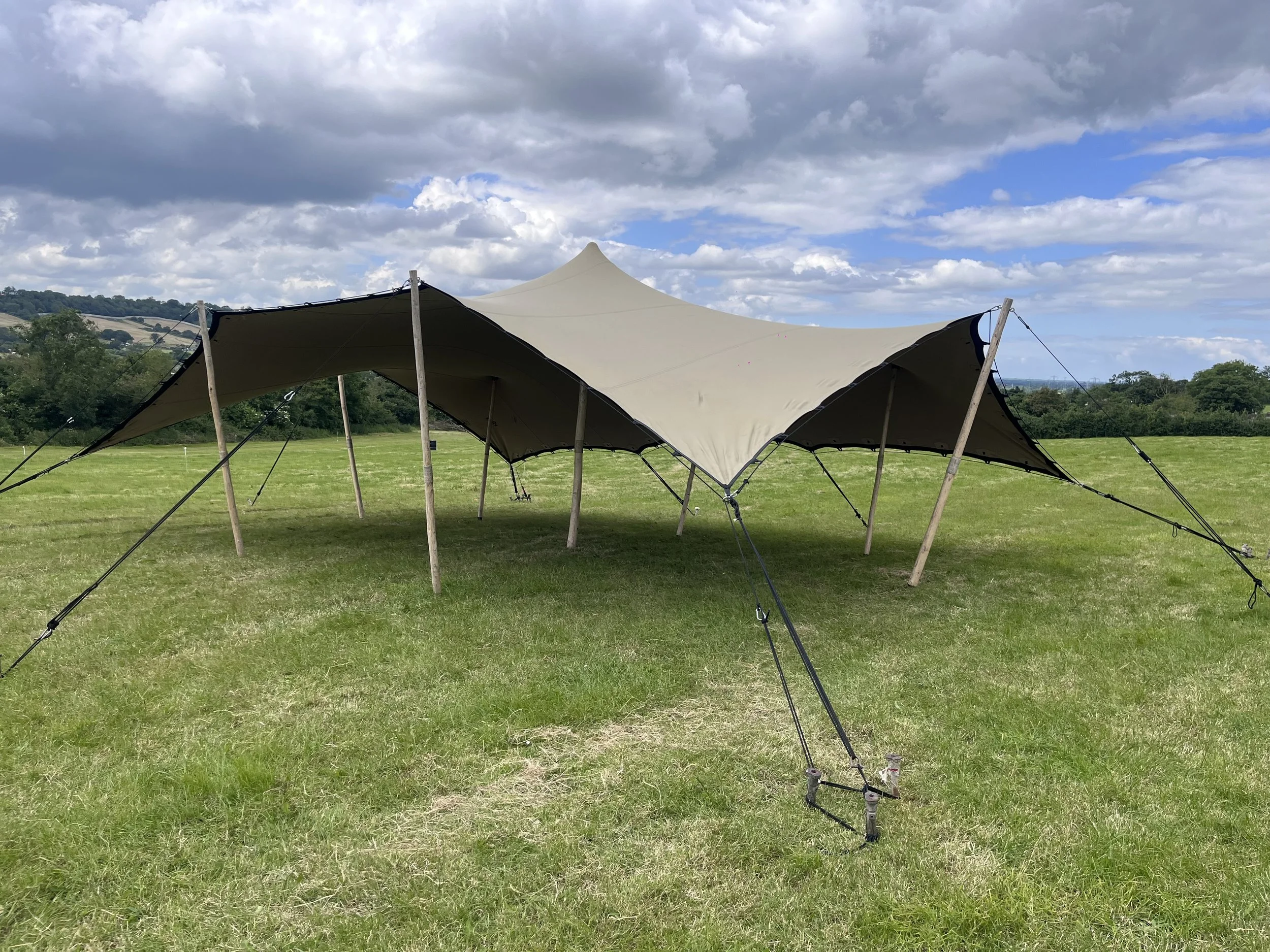 A large beige tent with black edges supported by wooden and metal poles, set up on a grassy field with rolling hills and trees in the background, under a partly cloudy sky.