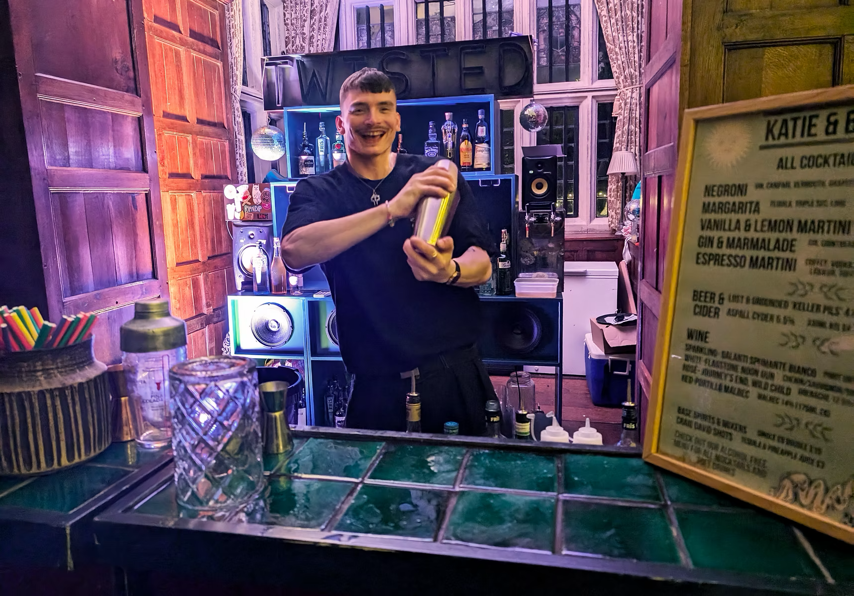 Happy man shaking a cocktail shaker behind a bar with colorful lighting, bottles of alcohol on shelves, a large window with curtains, and a menu on the right side.