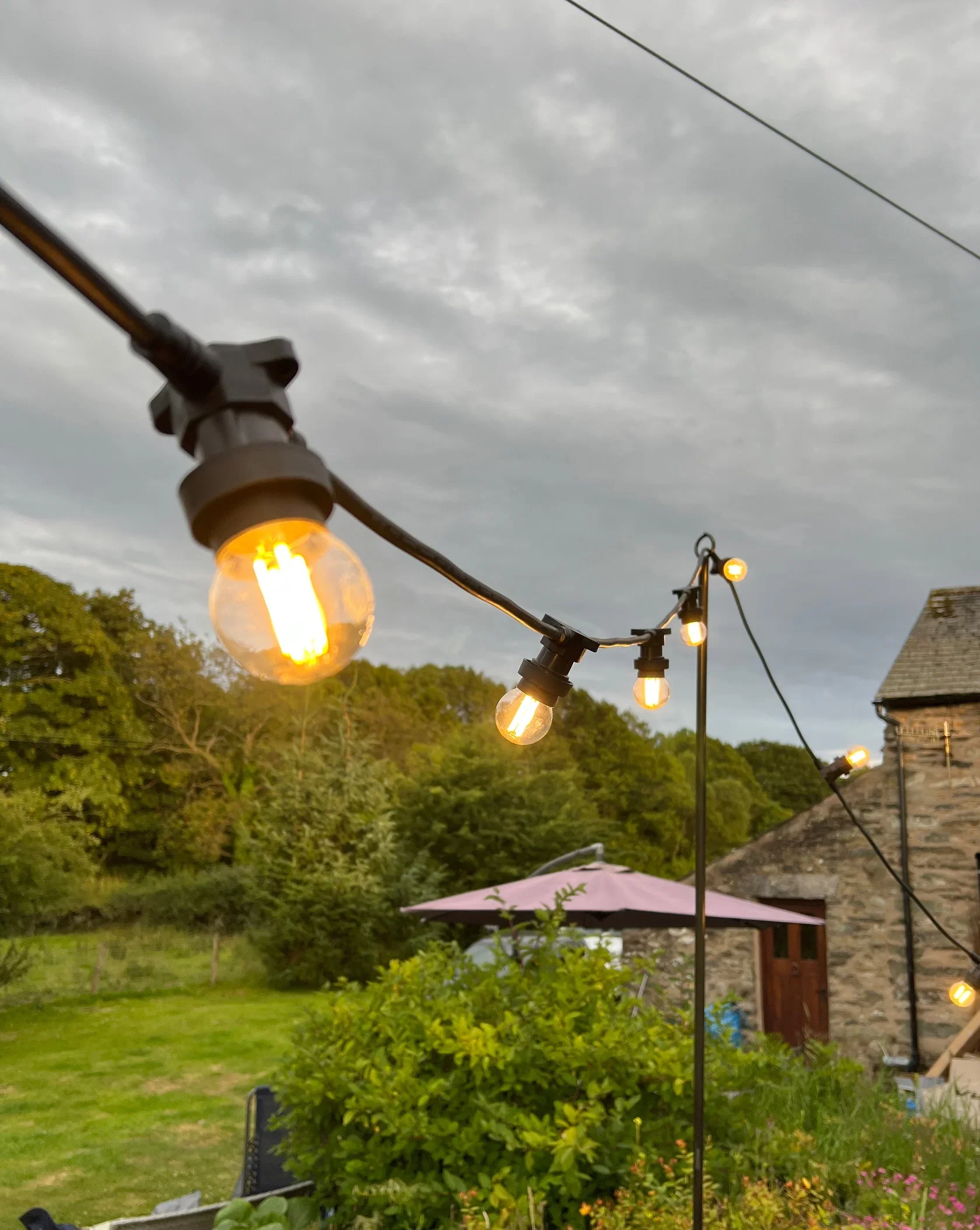 String of outdoor string lights hanging above a garden with trees, a stone building, a patio umbrella, and a cloudy sky.
