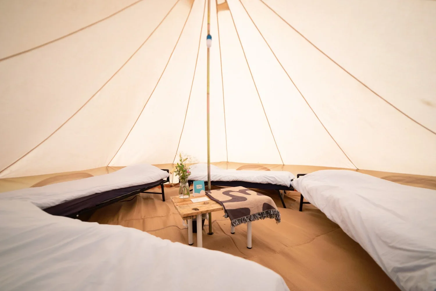 Inside a beige canvas teepee tent with four beds covered in white and dark linens, arranged in a semi-circle around a small rustic wooden table with a vase of flowers, a blue card, and a blanket.