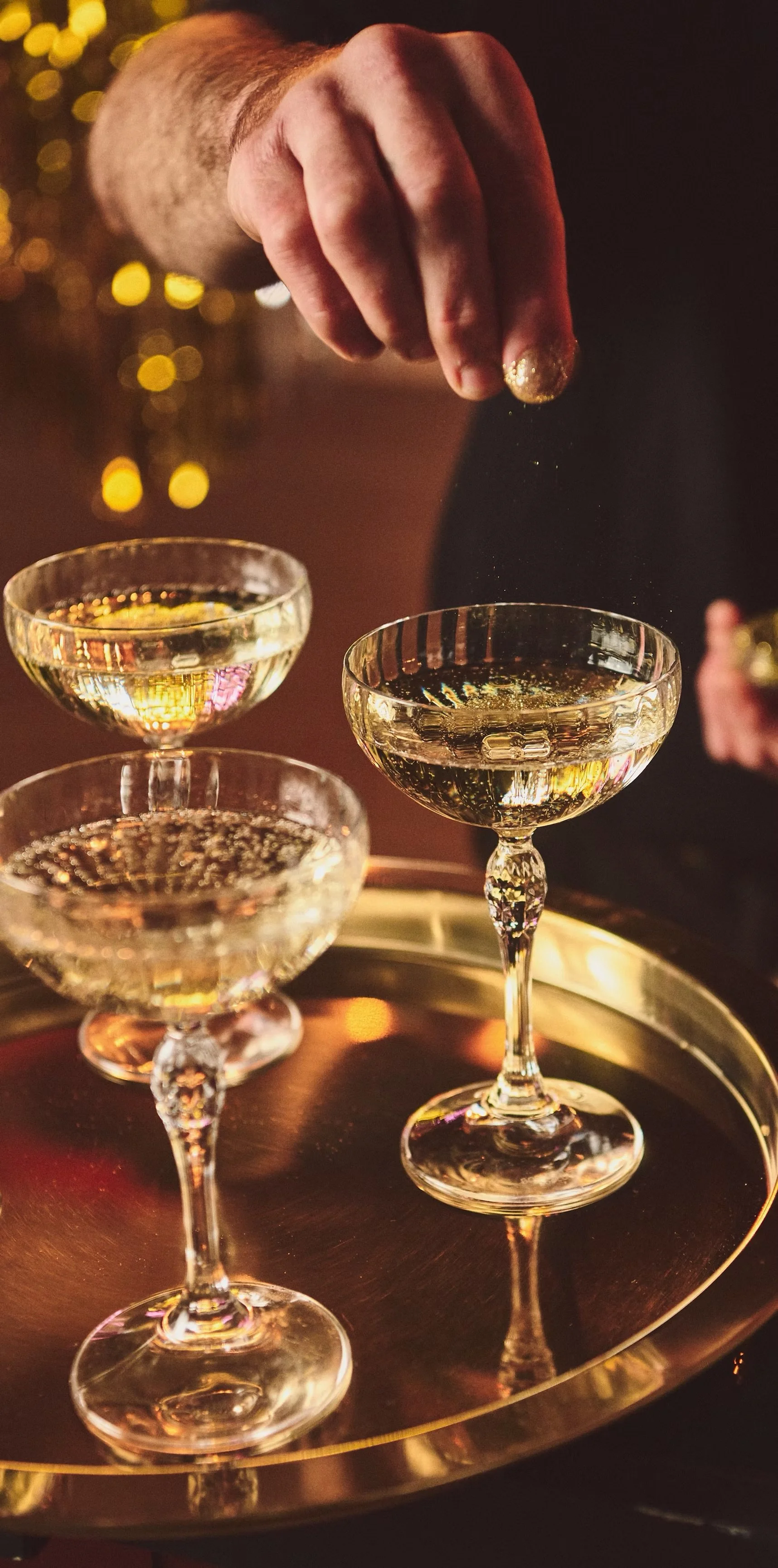 A hand sprinkling gold glitter onto champagne cocktails in coupe glasses on a gold tray.