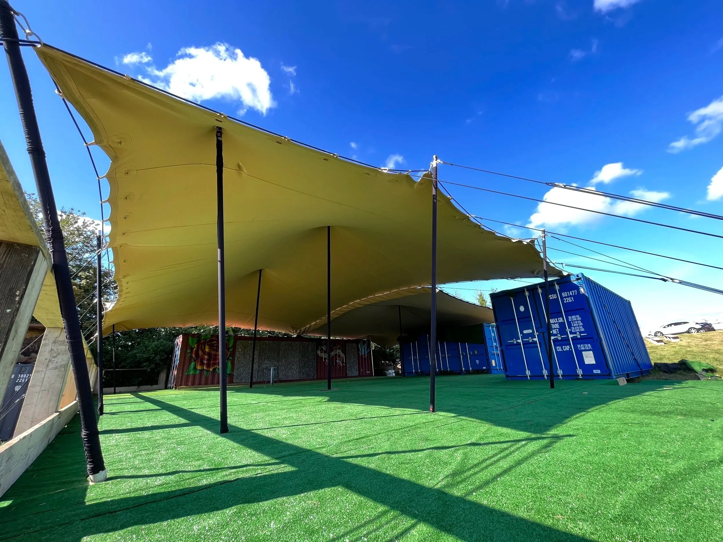 Outdoor stage with green artificial turf, large yellow canopy, and blue shipping container in the background under a partly cloudy sky.