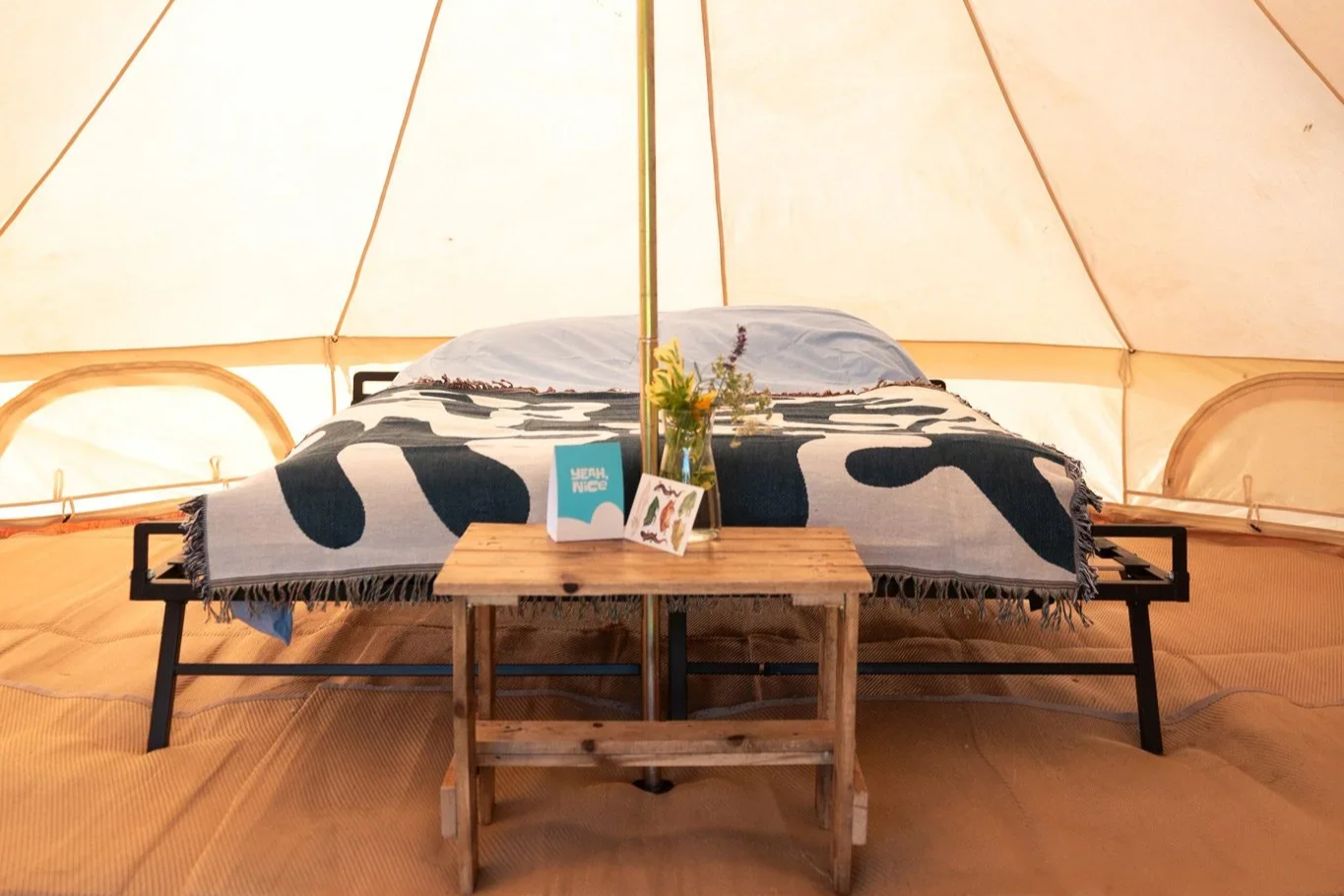 A bed inside a tent with a black metal frame, topped with a blanket featuring large black and white abstract shapes, and a pillow. A wooden table with flowers, cards, and a small sign are placed in front of the bed.