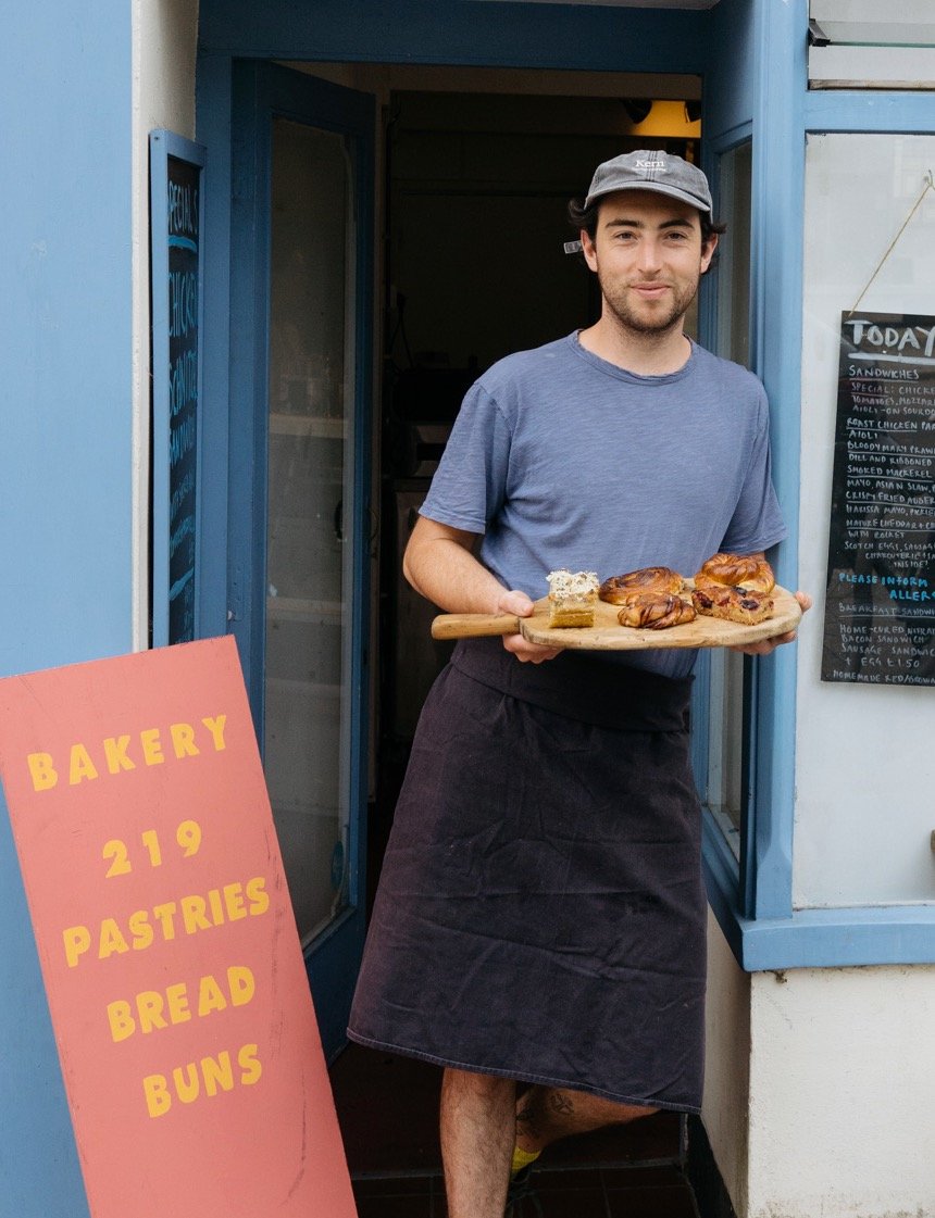 A young man standing at the entrance of a bakery holding a wooden tray with pastries. He is wearing a gray cap, a blue T-shirt, and a black apron. There is a pink sign with yellow text that reads 'Bakery 219 Pastries Bread Buns' and a chalkboard menu beside him.