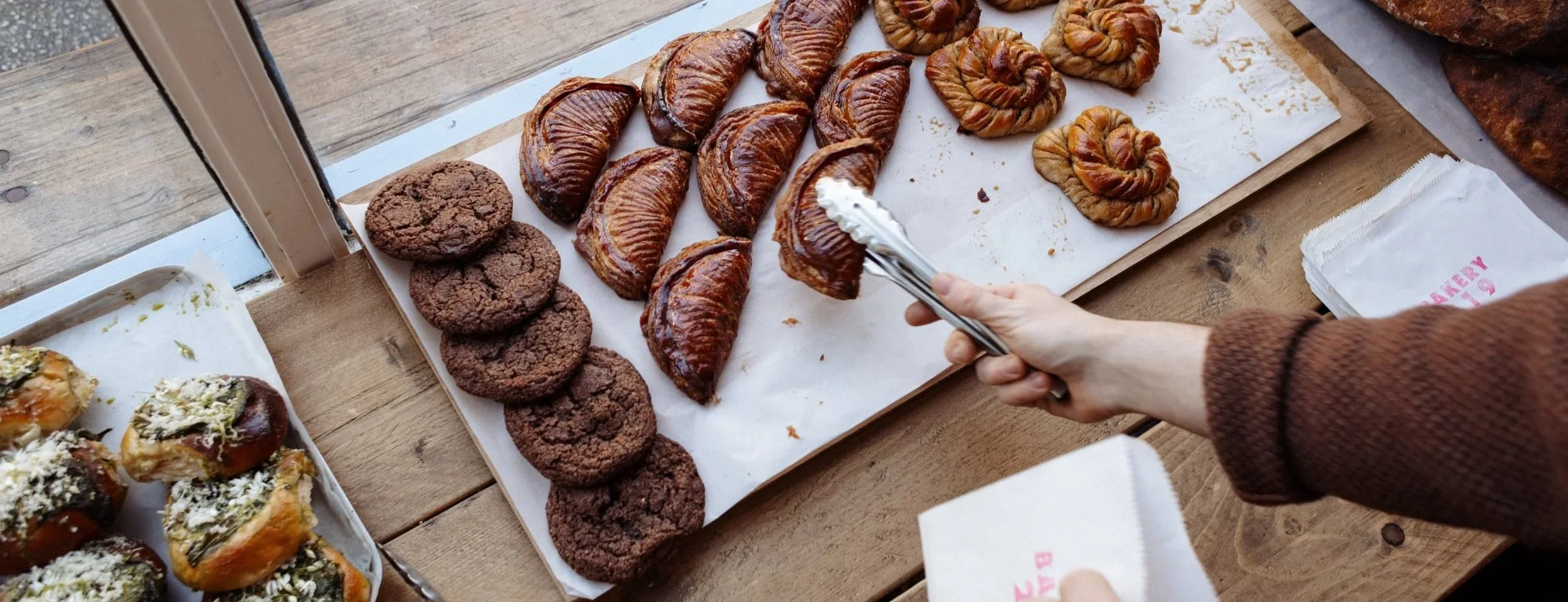 Person holding tongs near an assortment of baked pastries, including cookies, croissants, and Danish pastries, on a wooden table.