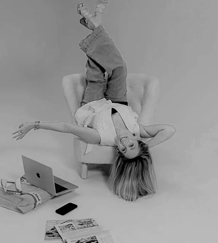 Brielle Fontaine smiling upside down in a white armchair beside her laptop in a minimalist studio.