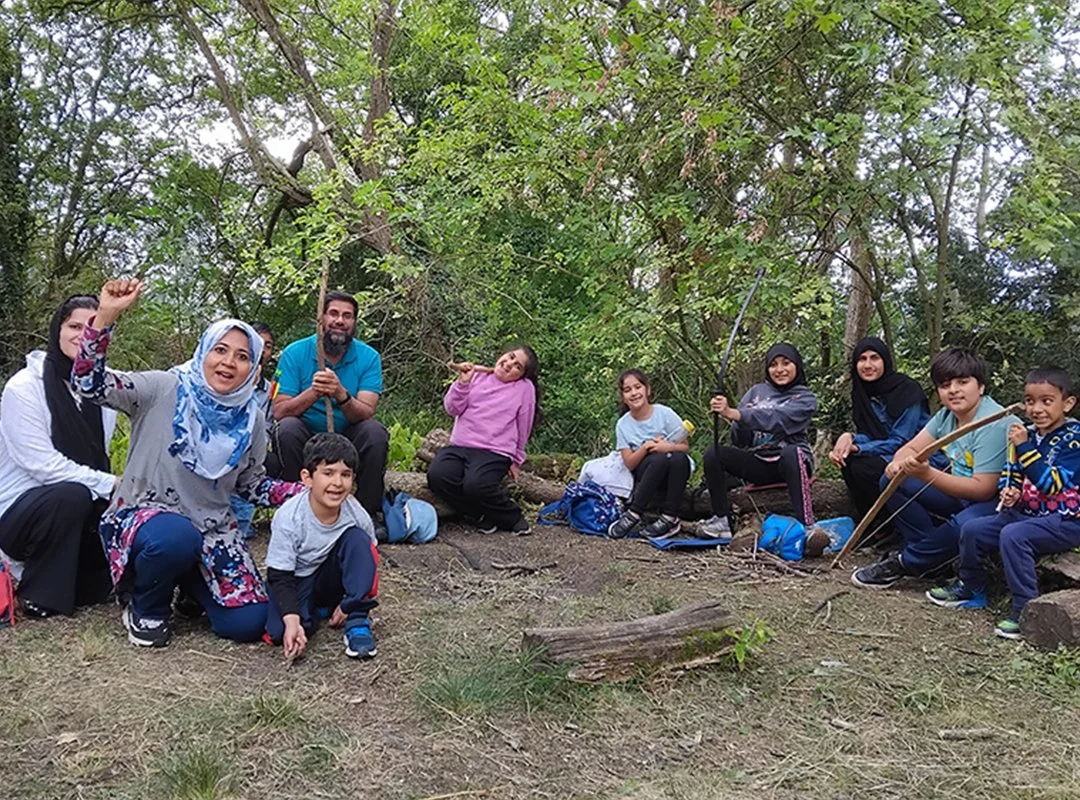 Group of children and adults outdoors in a wooded area, some holding sticks and a bow and arrow, posing for a photo.