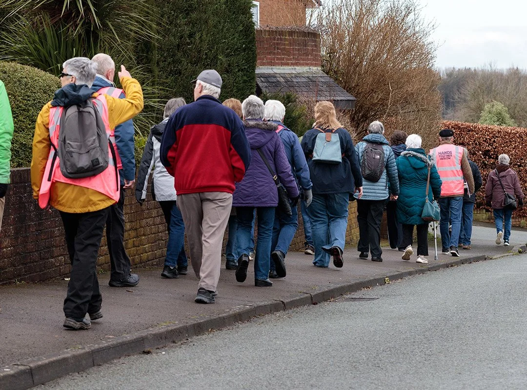 A group of elderly people walking together along a sidewalk in a suburban area, some wearing backpacks and jackets. One person wears a pink high-visibility vest.