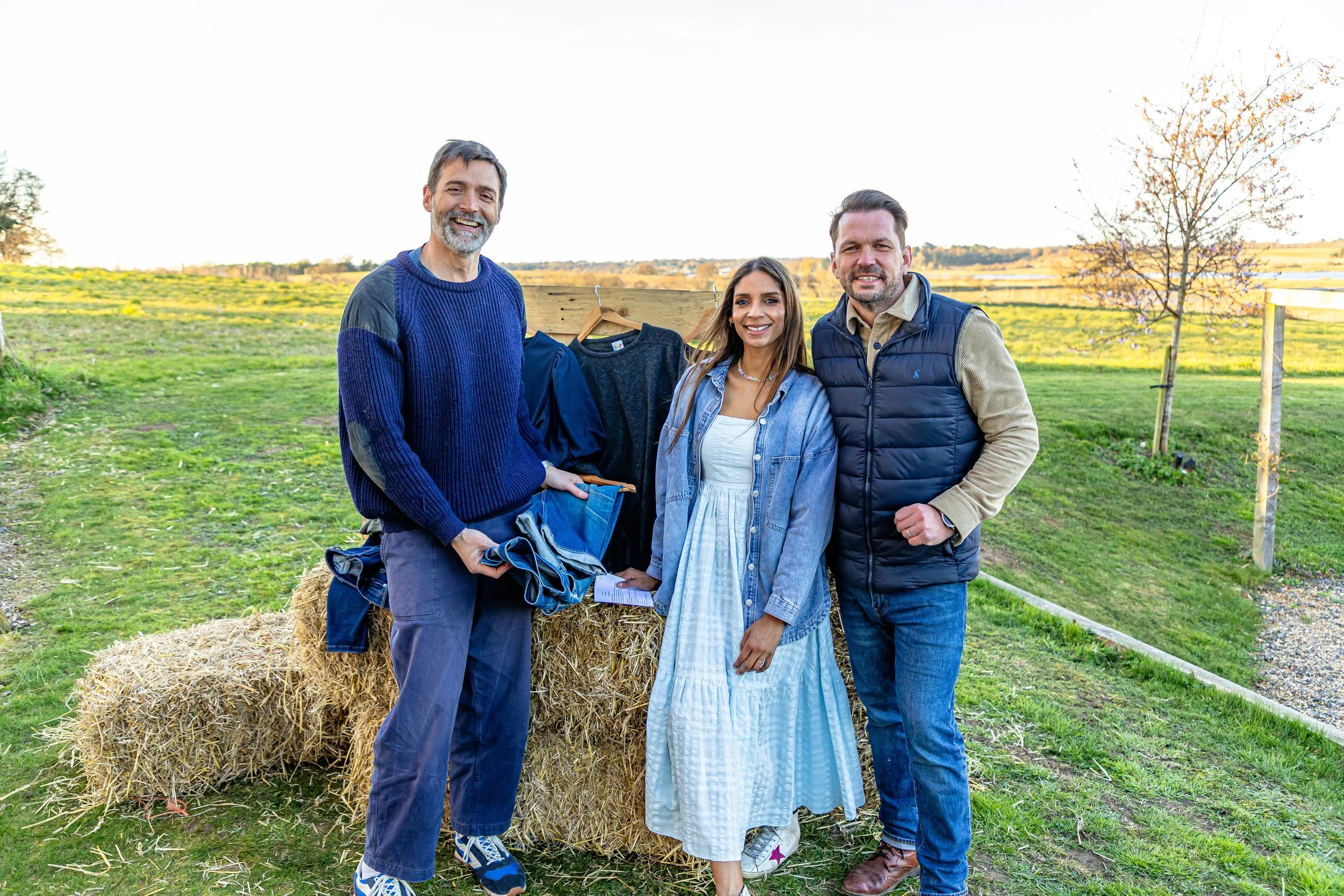 Three smiling people, two men and one woman, standing outdoors in a grassy field with a hay bale and a clothesline with hanging clothing behind them. They are dressed casually for mild weather, with a scenic rural landscape in the background and trees under a clear sky.