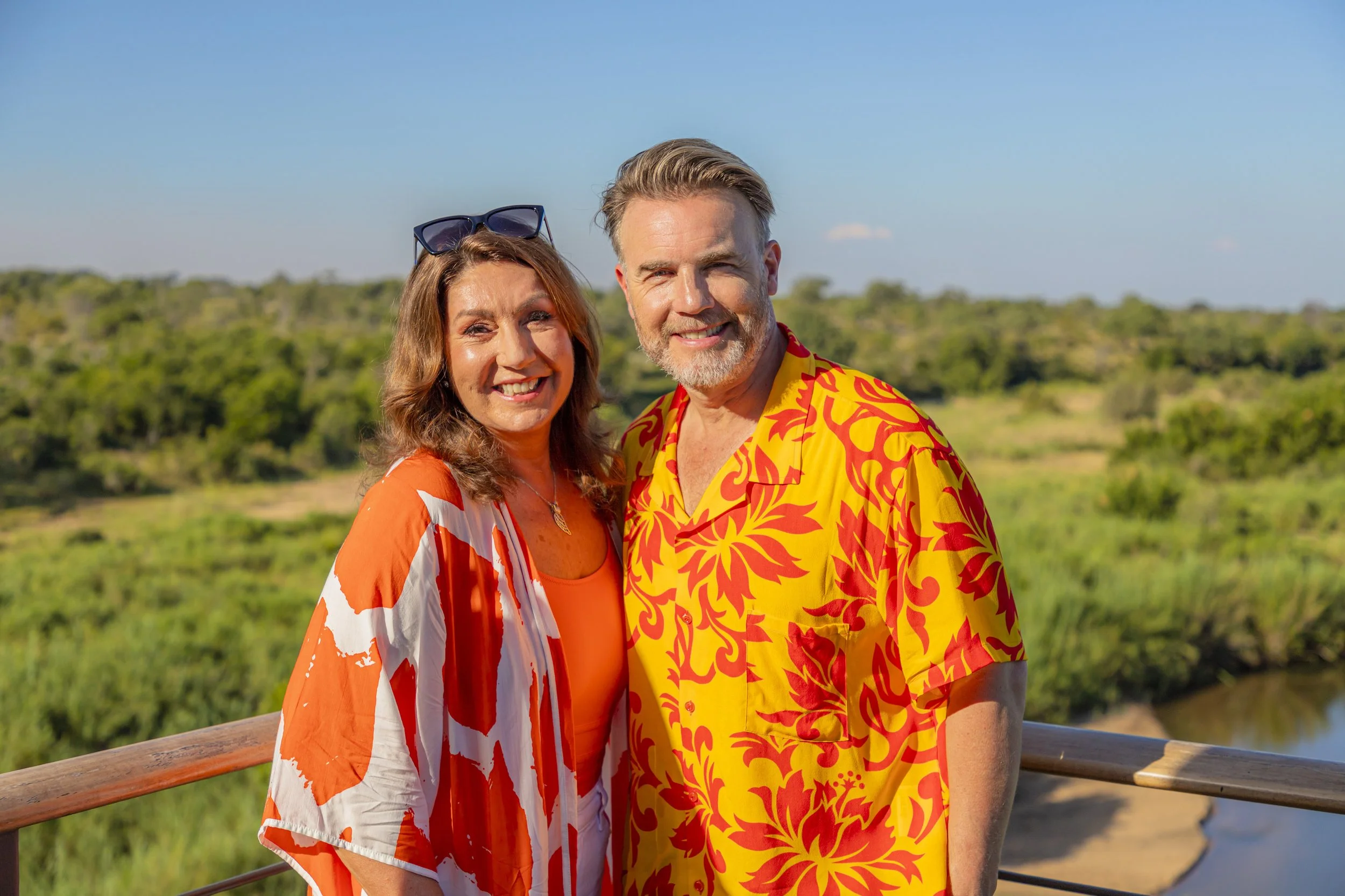 A smiling couple standing outdoors on a sunny day, wearing colorful tropical shirts, with a green landscape and blue sky in the background.