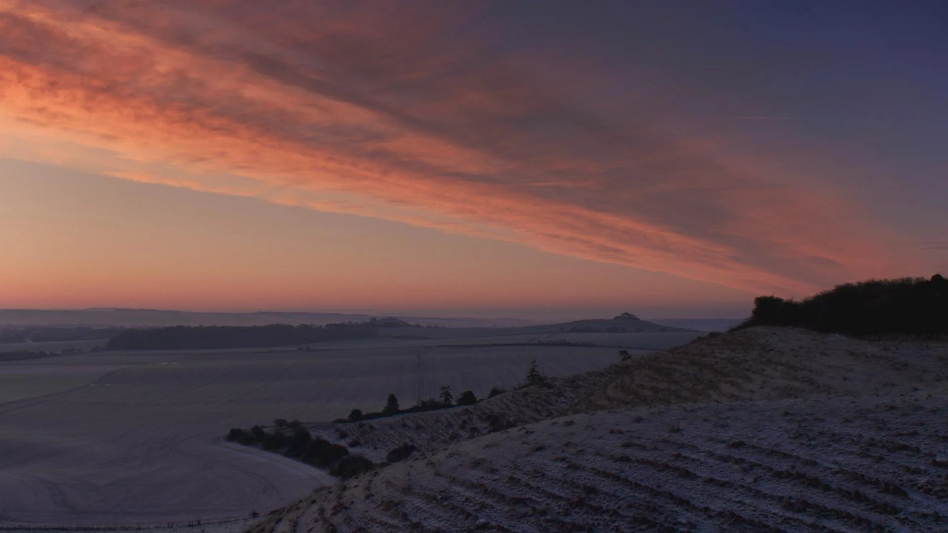 A colorful evening sky with pink, orange, and purple hues above rolling hills and a landscape with sparse trees.