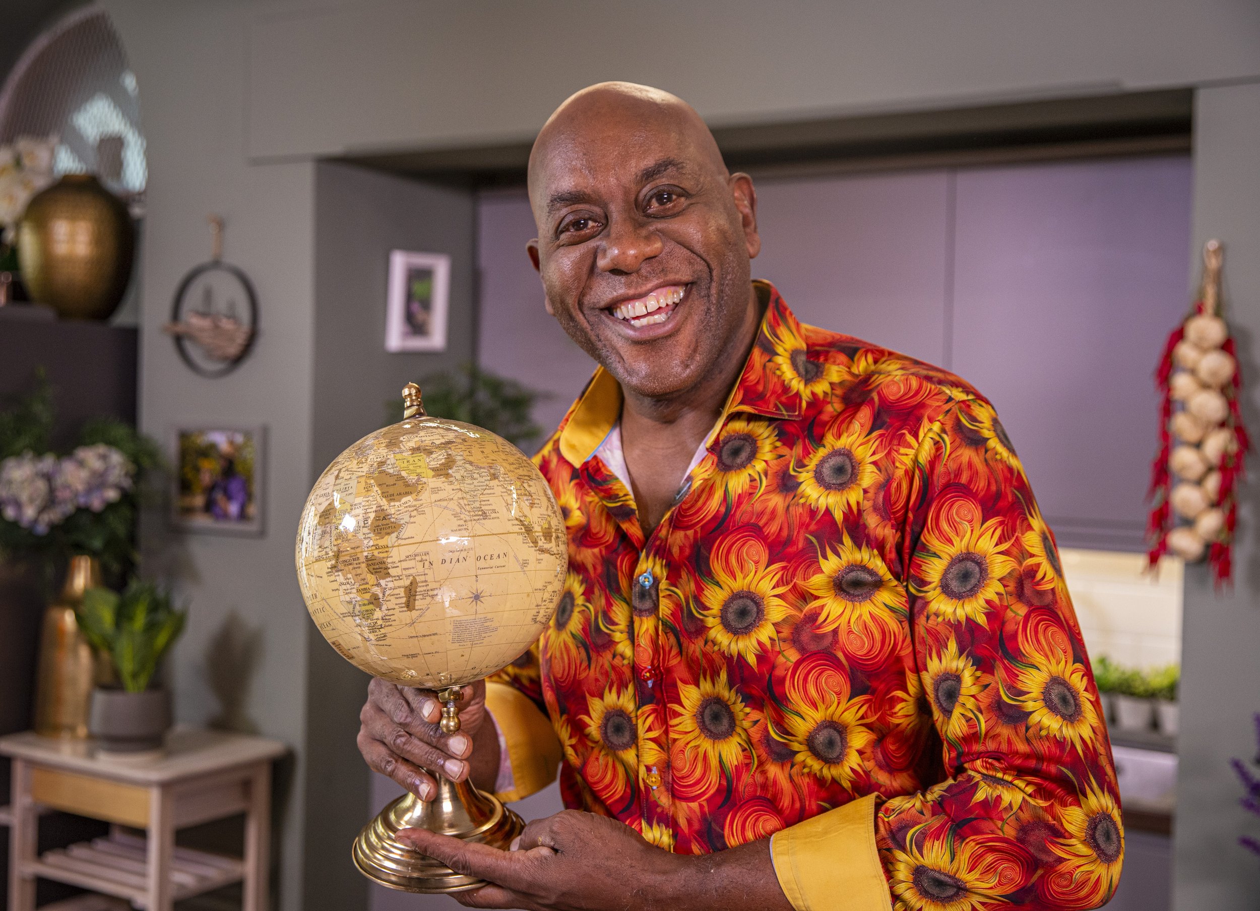 A smiling man with a bald head wearing a vibrant sunflower-patterned shirt holding a globe in an indoor setting decorated with plants and framed pictures.