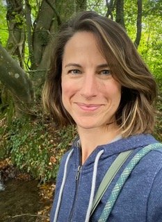 A woman outdoors near a wooded area with green trees in the background, smiling and wearing a blue hoodie.