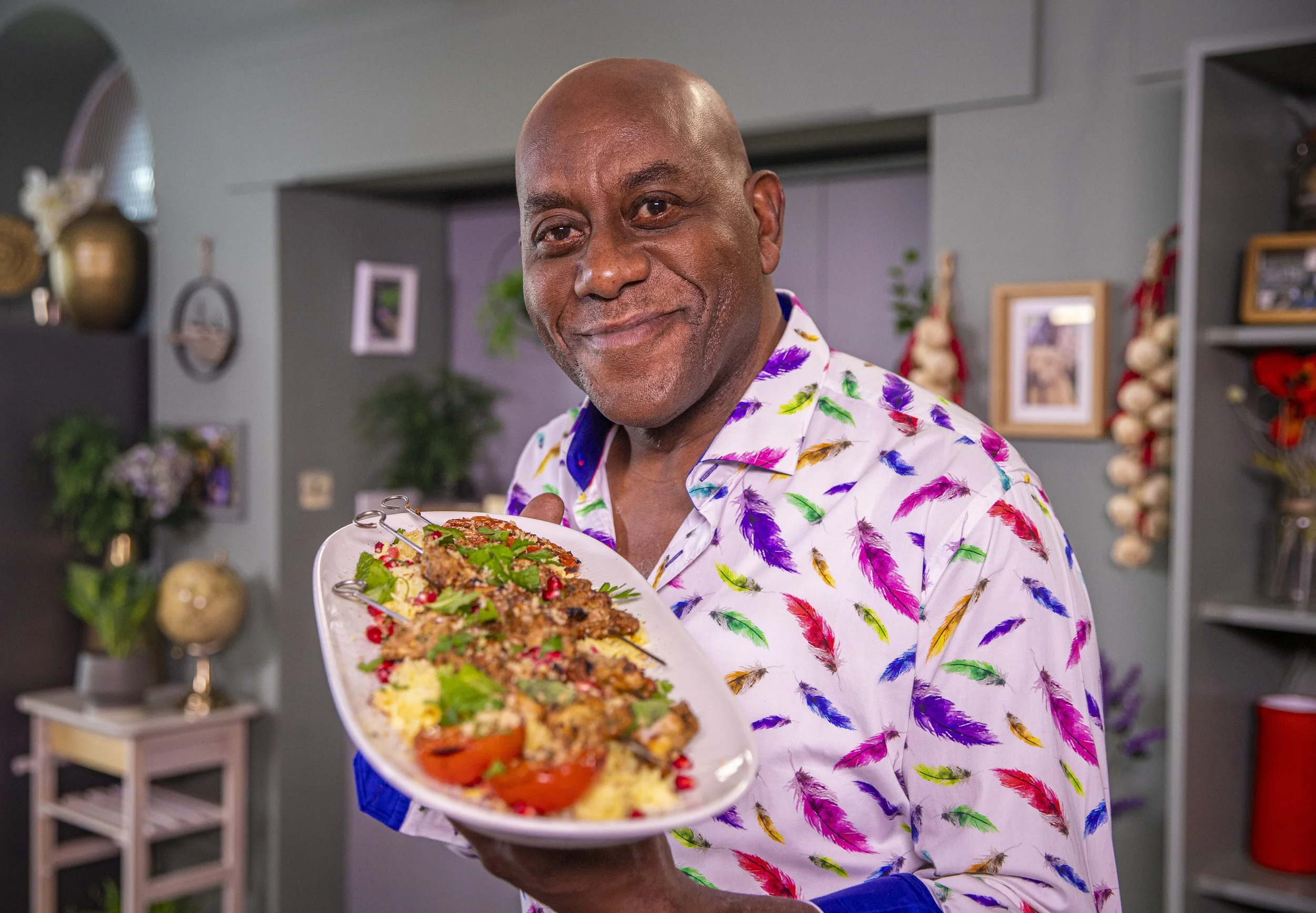 A man in a colorful feather-patterned shirt holding a platter of food, smiling in a decorated room.