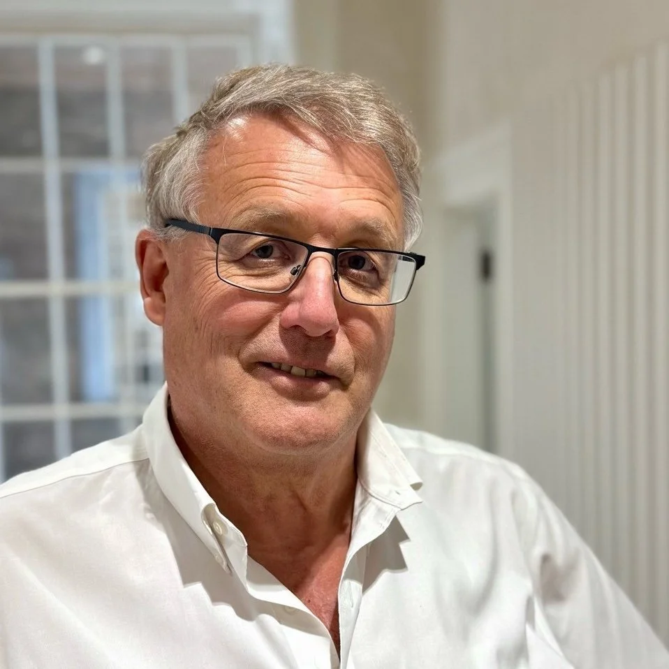 A middle-aged man with gray hair and glasses smiling, wearing a white collared shirt. He is indoors with a blurred background showing windows and a radiator.
