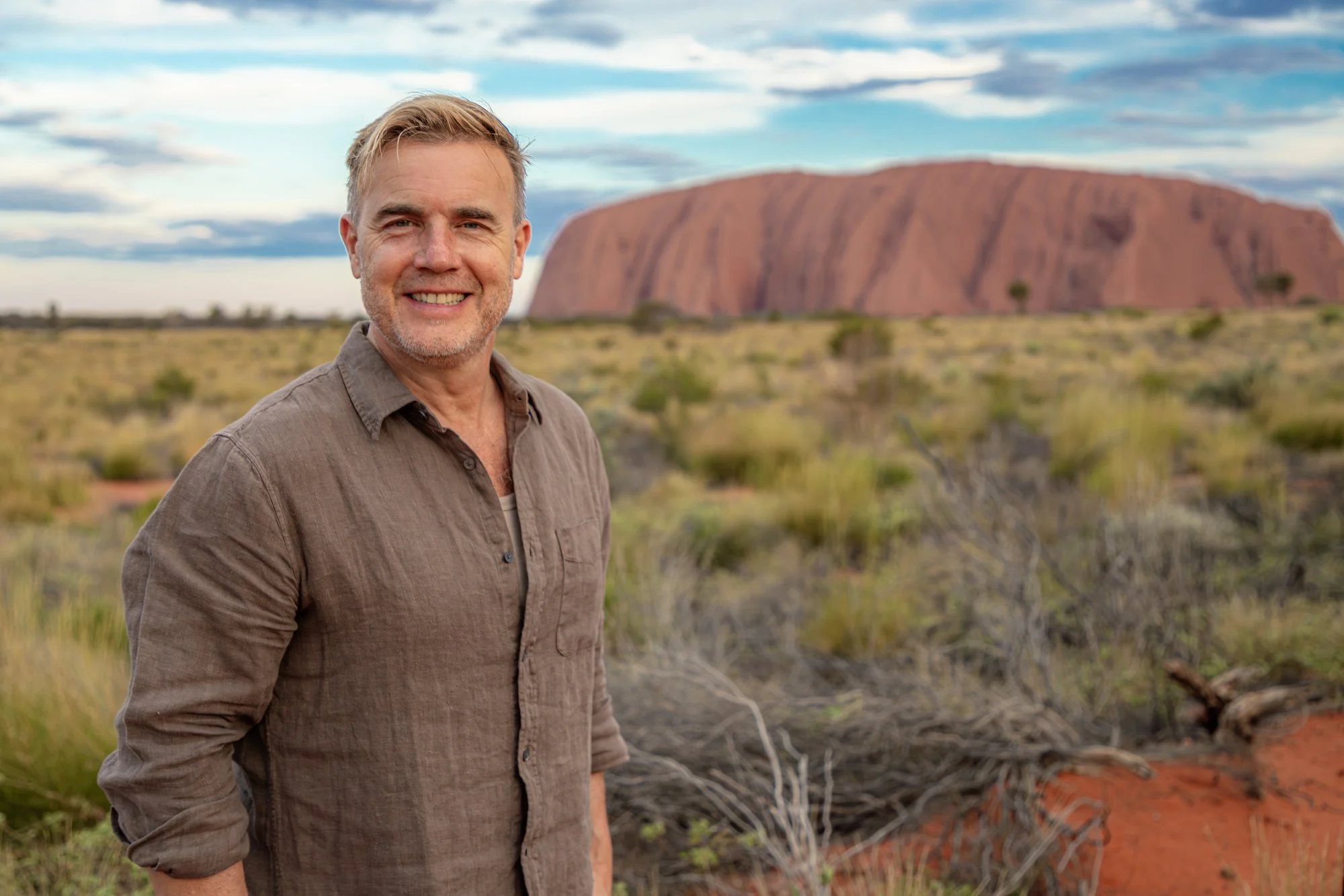 A man smiling at the camera in a semi-arid landscape with the Uluru rock formation in the background during daytime