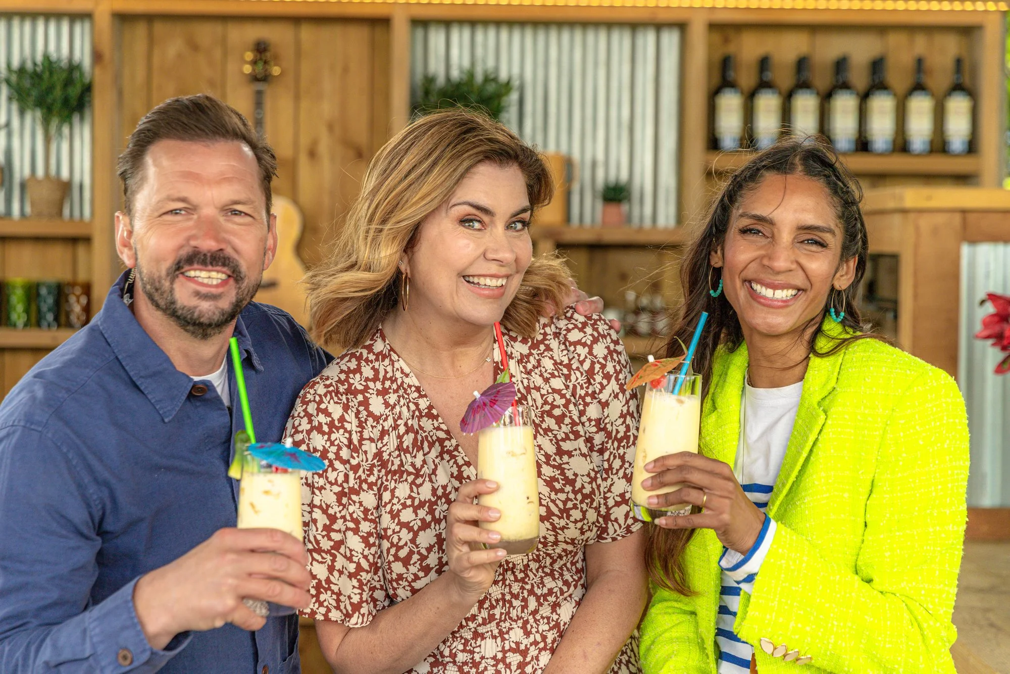 Three people smiling and holding tropical drinks with umbrellas and straws at a social gathering indoors.
