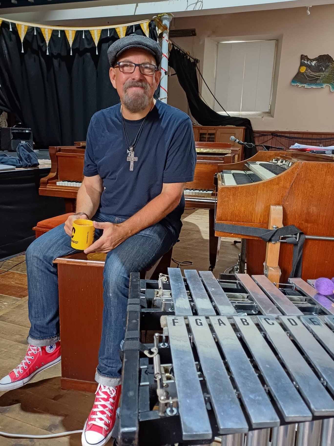 Man with glasses, beard, and a cap sitting in a music studio, holding a yellow cup, surrounded by musical instruments including a xylophone in the foreground and pianos in the background.