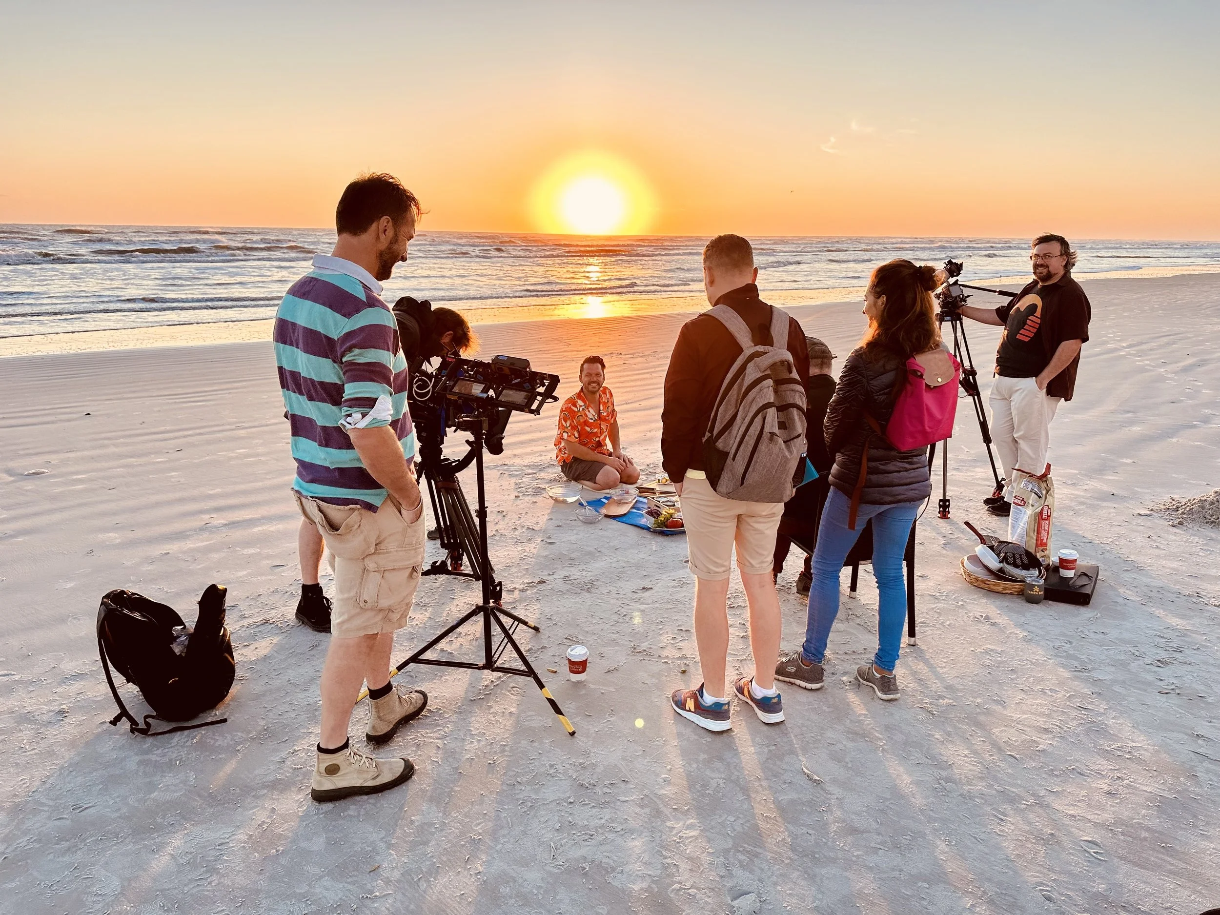 A film crew shoots a scene on a beach at sunset, with one actor kneeling on the sand and surrounded by cameras, lights, and crew members.