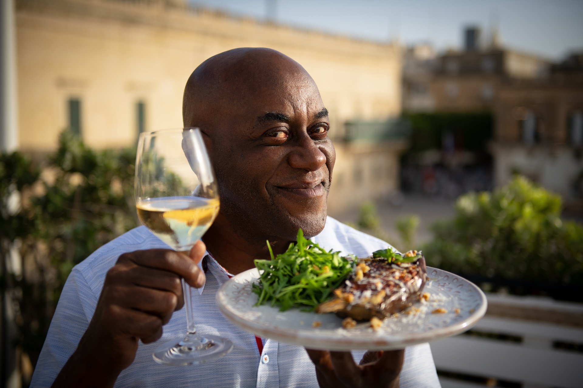 A man with a bald head and dark skin holding a glass of white wine in one hand and a plate with salad and grilled meat in the other, smiling outdoors with blurred buildings in the background.