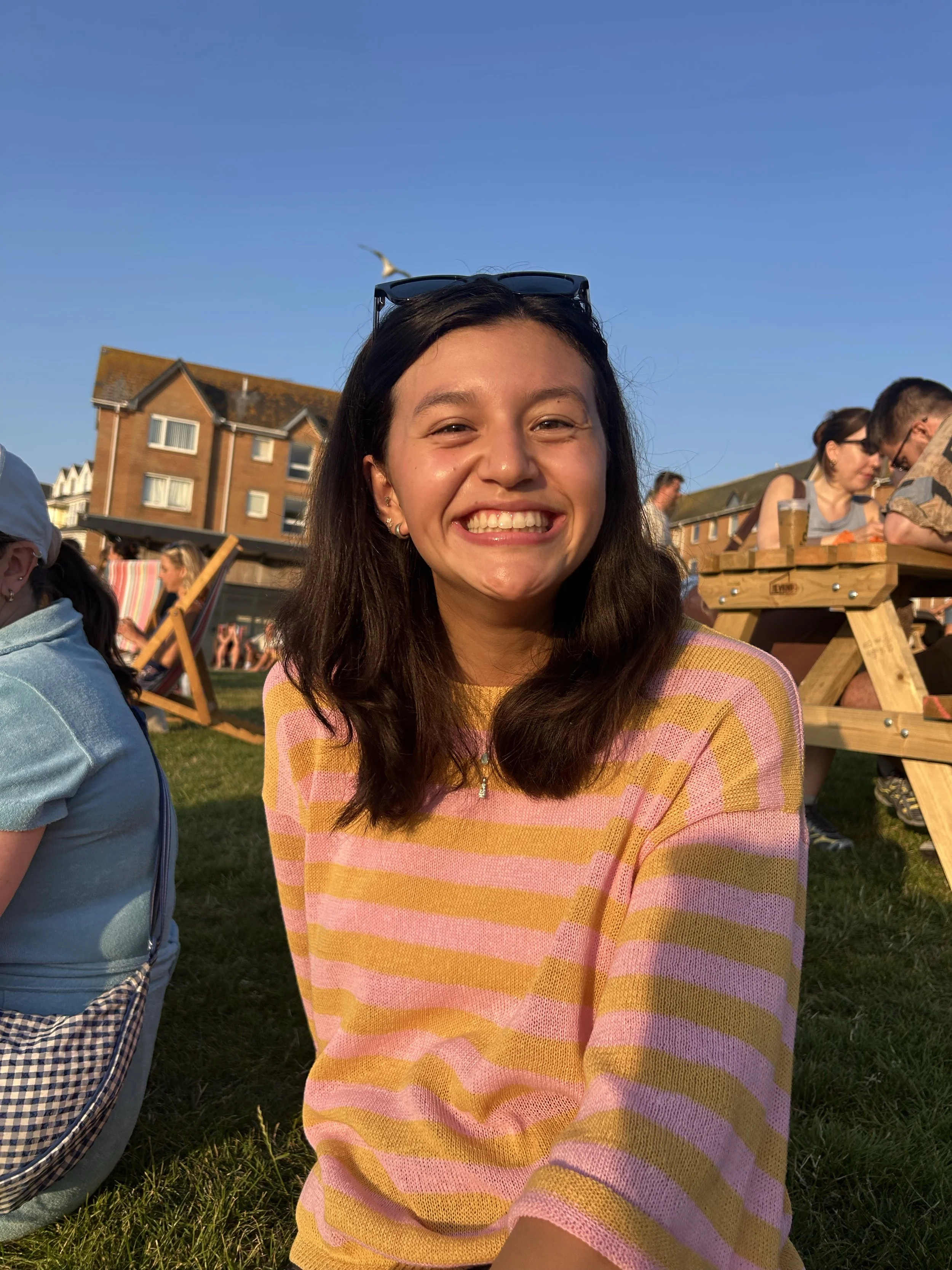 A young woman with long dark hair, smiling widely, wearing sunglasses on her head, and a pastel-colored striped sweater, outdoors during the daytime with a clear blue sky, surrounded by other people and wooden picnic tables.