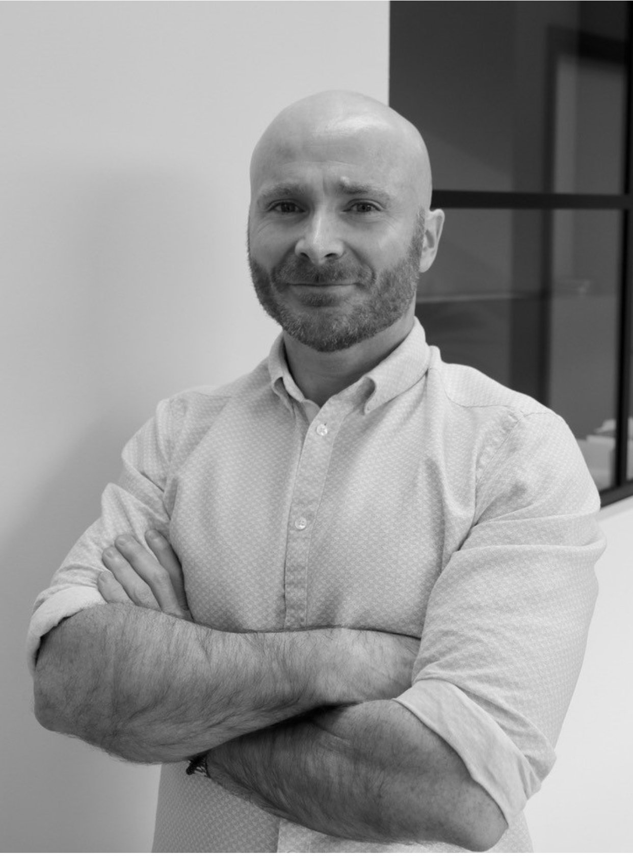 Black and white portrait of a man with a beard, bald head, wearing a button-up shirt, standing with arms crossed in front of a plain wall and a window.