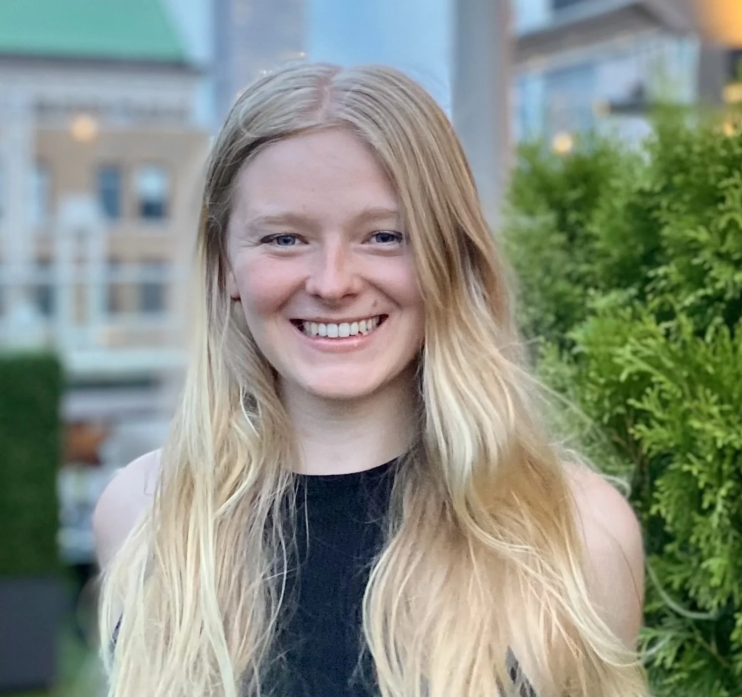 Portrait of a smiling woman with long blonde hair, wearing a black top, standing outdoors with a blurred background of buildings and greenery.