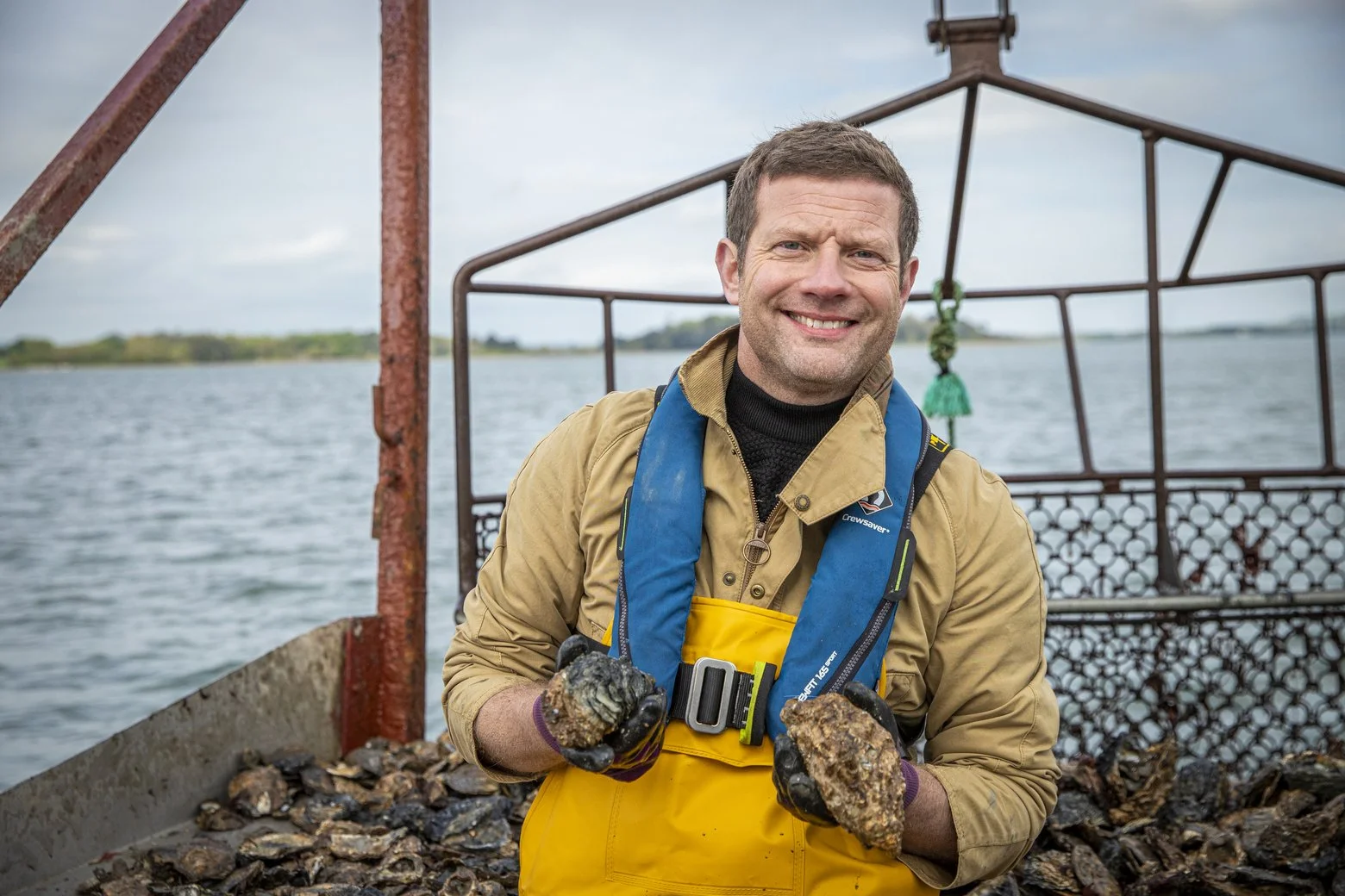 A man in yellow waterproof gear holding oysters on a boat by a body of water, smiling near a metal structure and a pile of oysters.