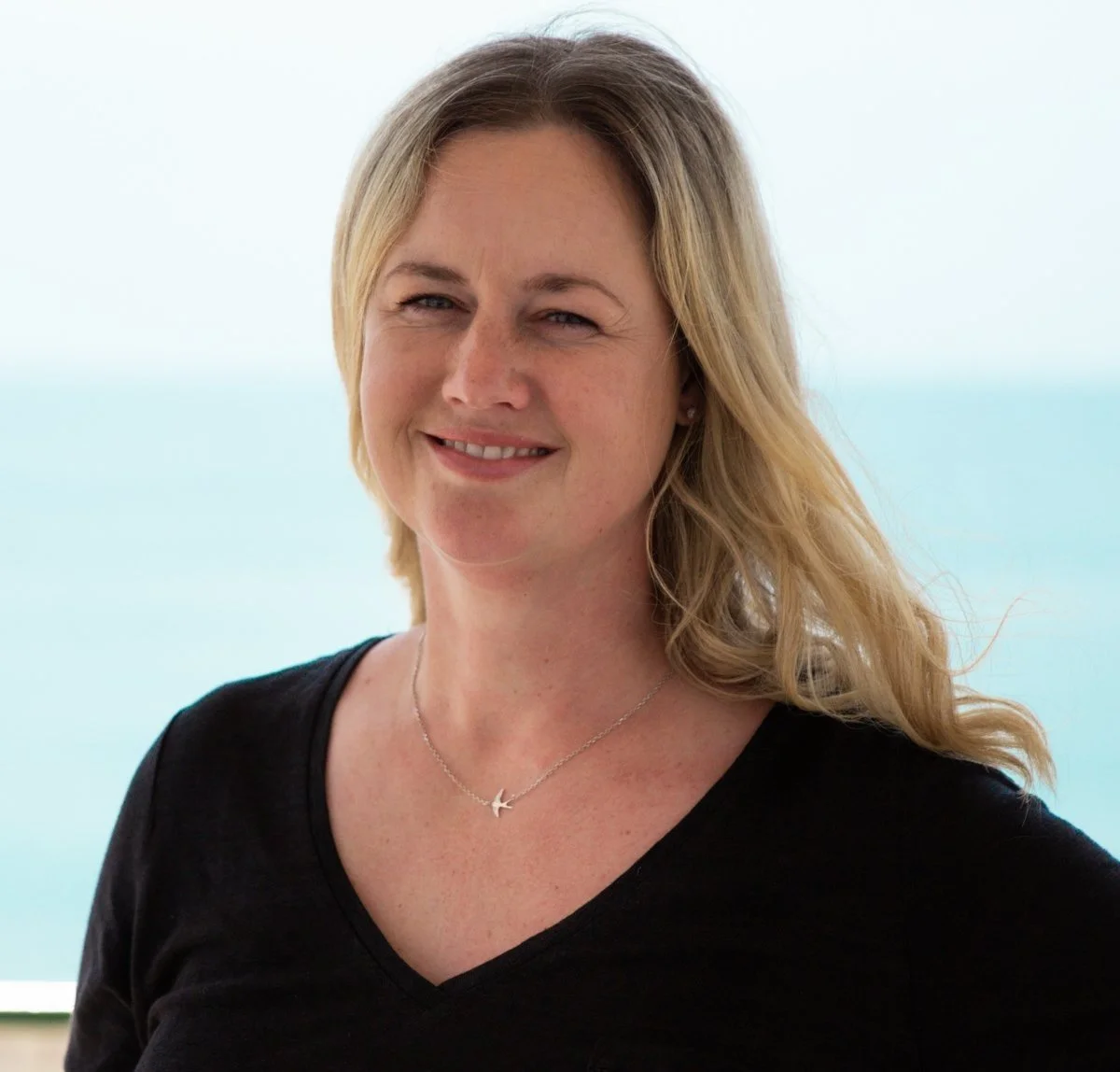 A woman with blonde hair smiling, wearing a black top and a silver bird necklace, against a background of the ocean and sky.