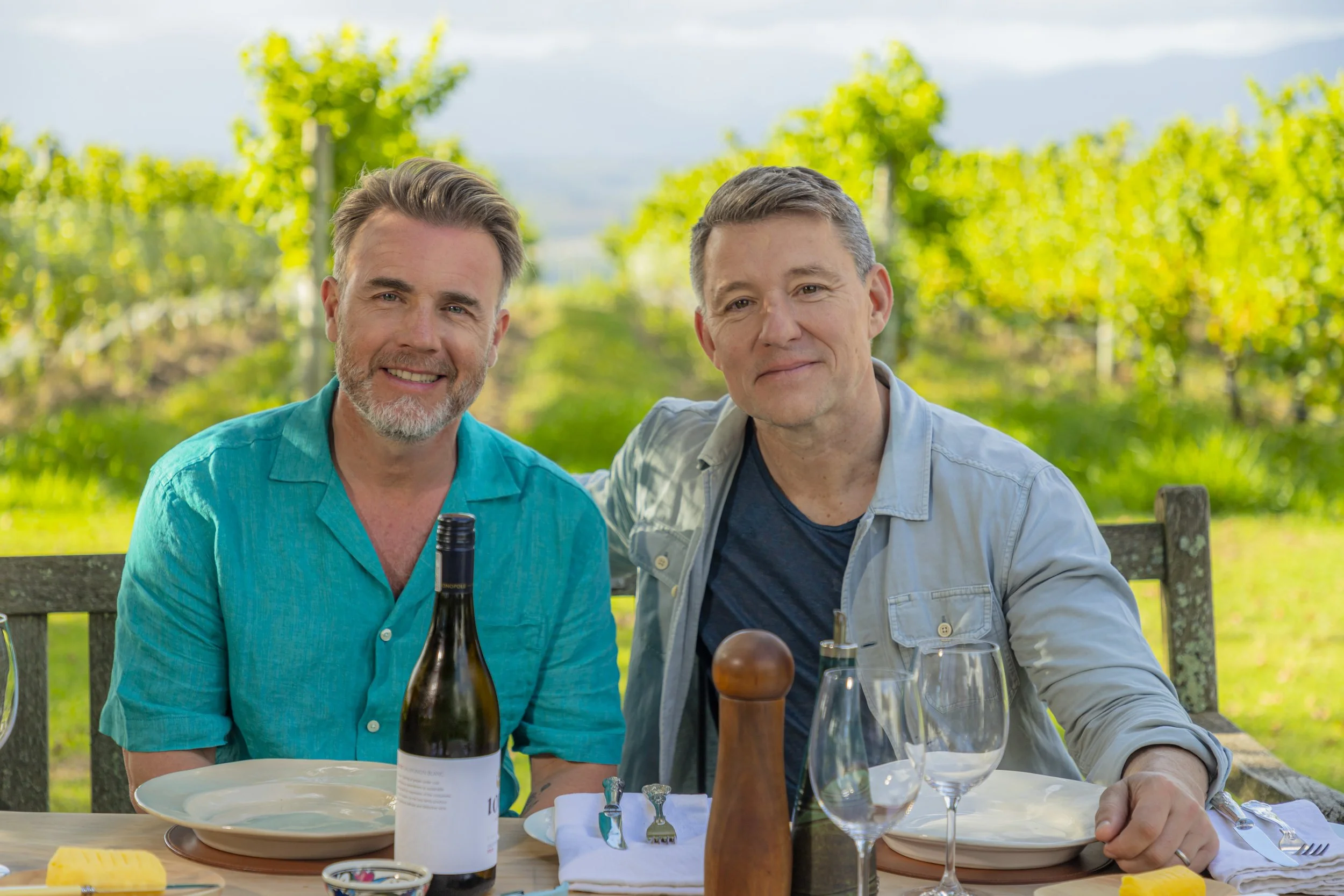Two middle-aged men sitting at an outdoor table with plates, wine, and condiments, smiling at the camera, with a vineyard background on a sunny day.