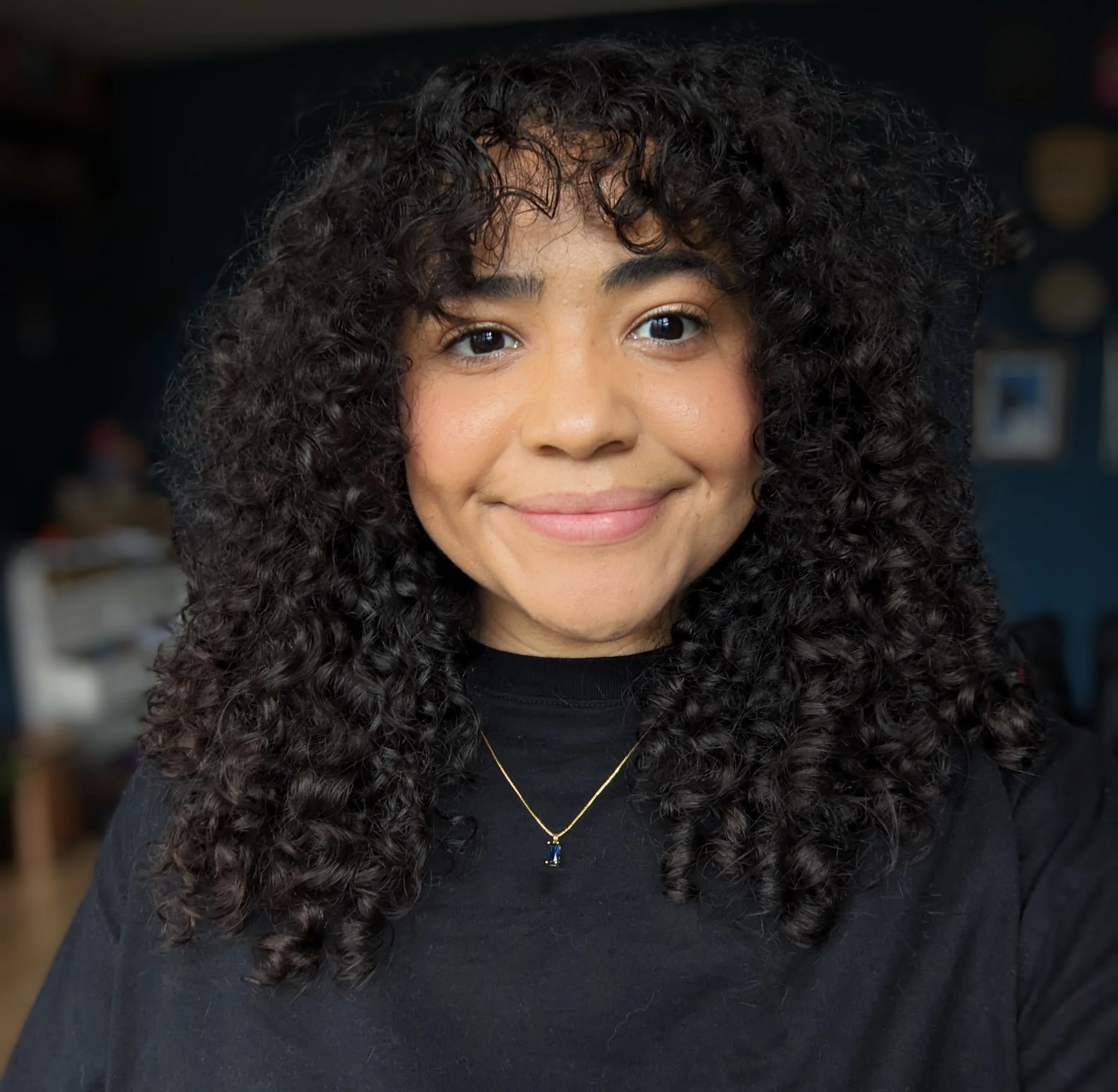 Close-up of a smiling woman with curly dark hair, wearing a black top and a delicate gold necklace with a pendant.