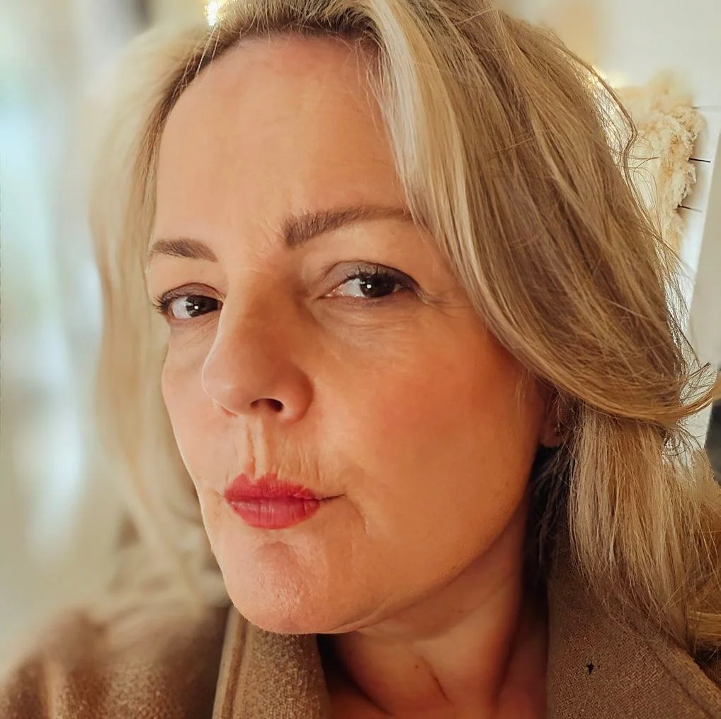 Close-up of a woman with blonde, wavy hair, wearing makeup, looking at the camera with a soft expression.