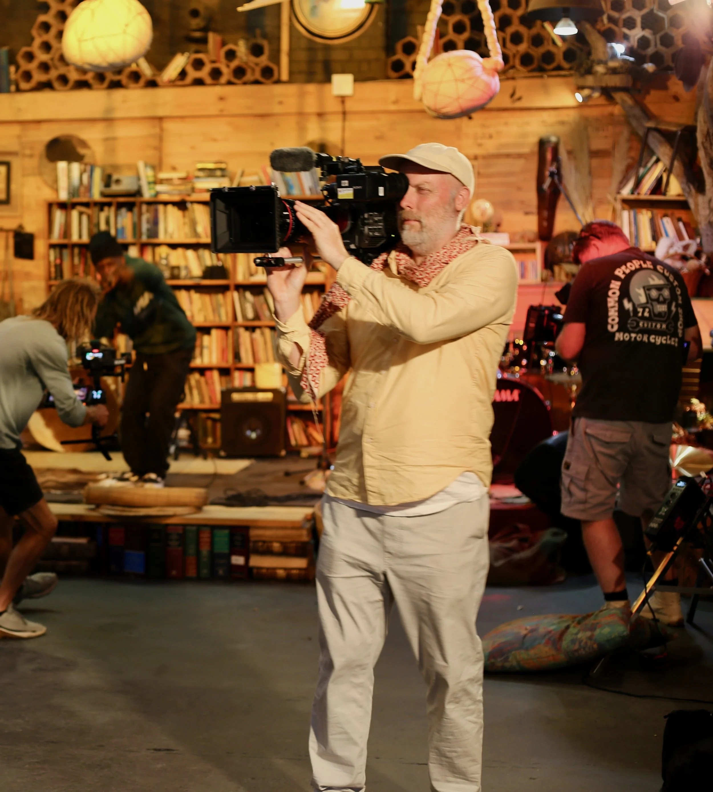 A man wearing a beige cap, a beige jacket, and white pants is holding a professional video camera and filming. He is inside a cozy room with wooden walls, bookshelves filled with books, and hanging paper lanterns. People are visible in the background, some working on equipment or engaging with each other.