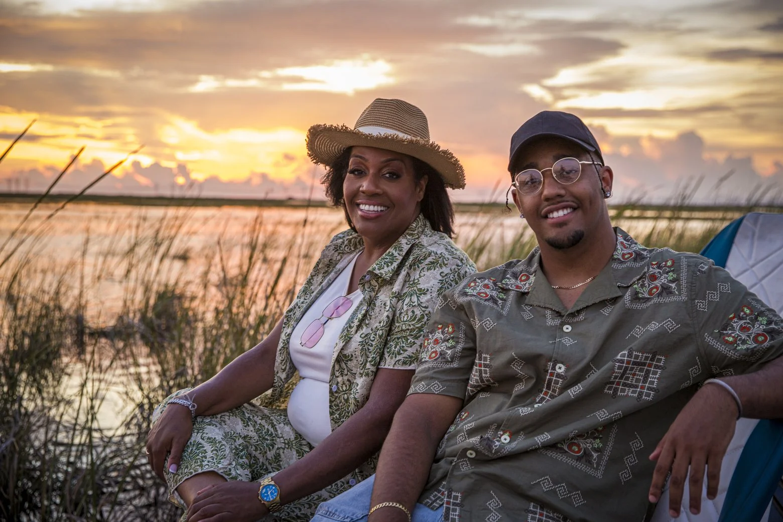 A smiling woman and man sitting outdoors during sunset near a body of water, with tall grass in the foreground.