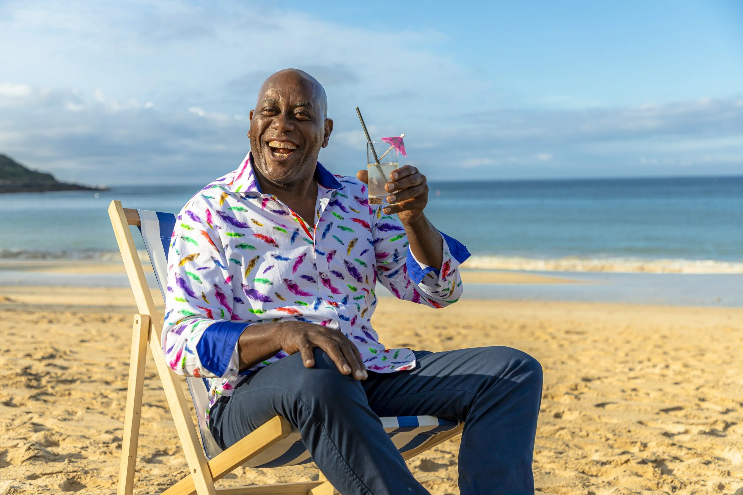 A happy man wearing a colorful, patterned shirt sits on a beach chair, smiling and holding a drink with a small pink umbrella, against a backdrop of the ocean and a partly cloudy sky.