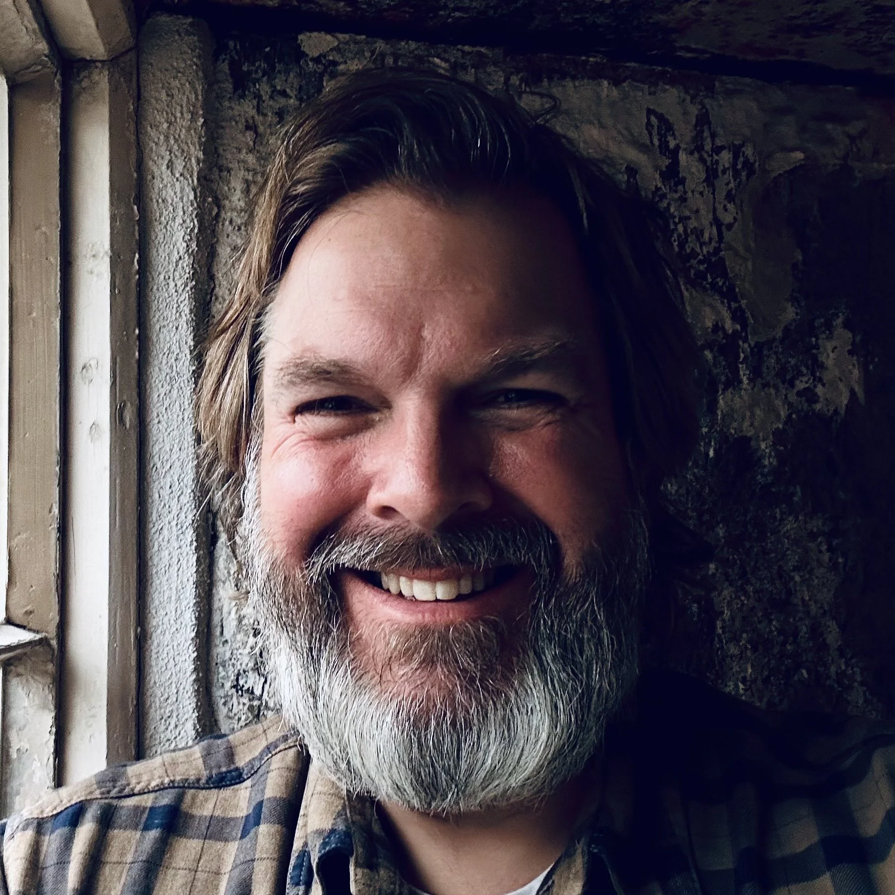 Partial close-up of smiling man with long hair, beard, mustache, wearing plaid shirt, standing near a stone wall.