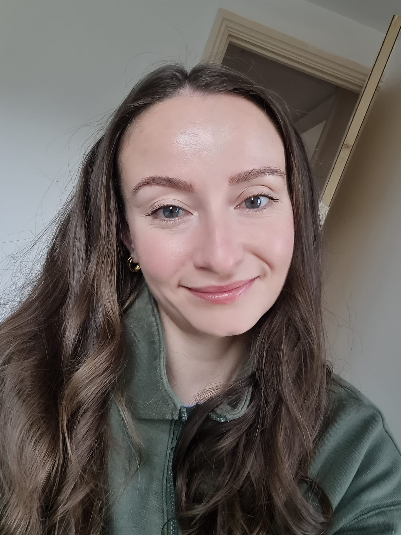 A woman with long brown hair and blue eyes smiling, wearing a green jacket, in an indoor setting with a door in the background.