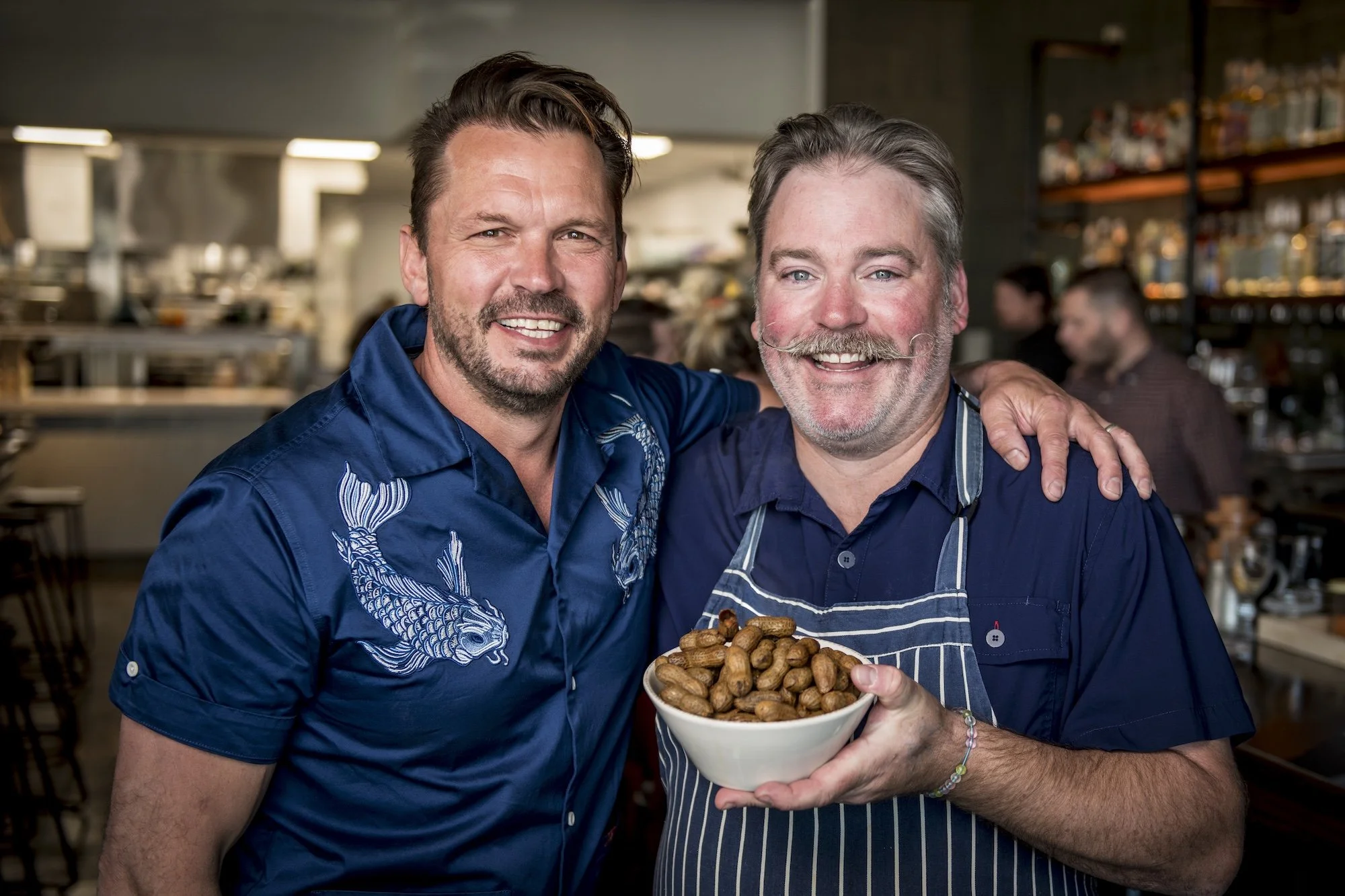 Two smiling men in blue shirts, one with a floral pattern and the other with an apron, are holding a bowl of peanuts together in a busy kitchen or restaurant setting.