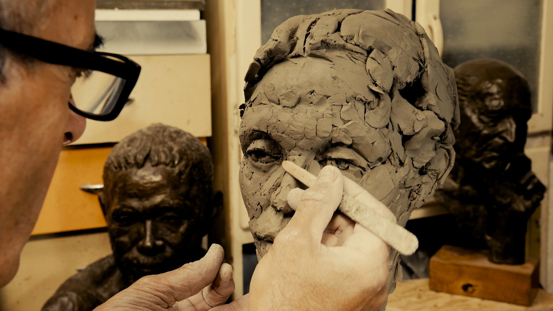 An artist working on a clay bust sculpture, shaping the facial features with a tool, surrounded by other bust sculptures in a workshop.